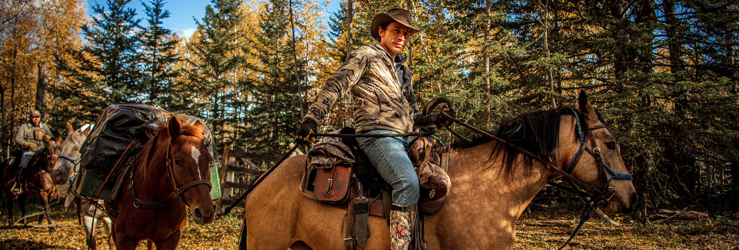 Cowboy riding a horse through an autumn forest with pack horses and a second rider behind him