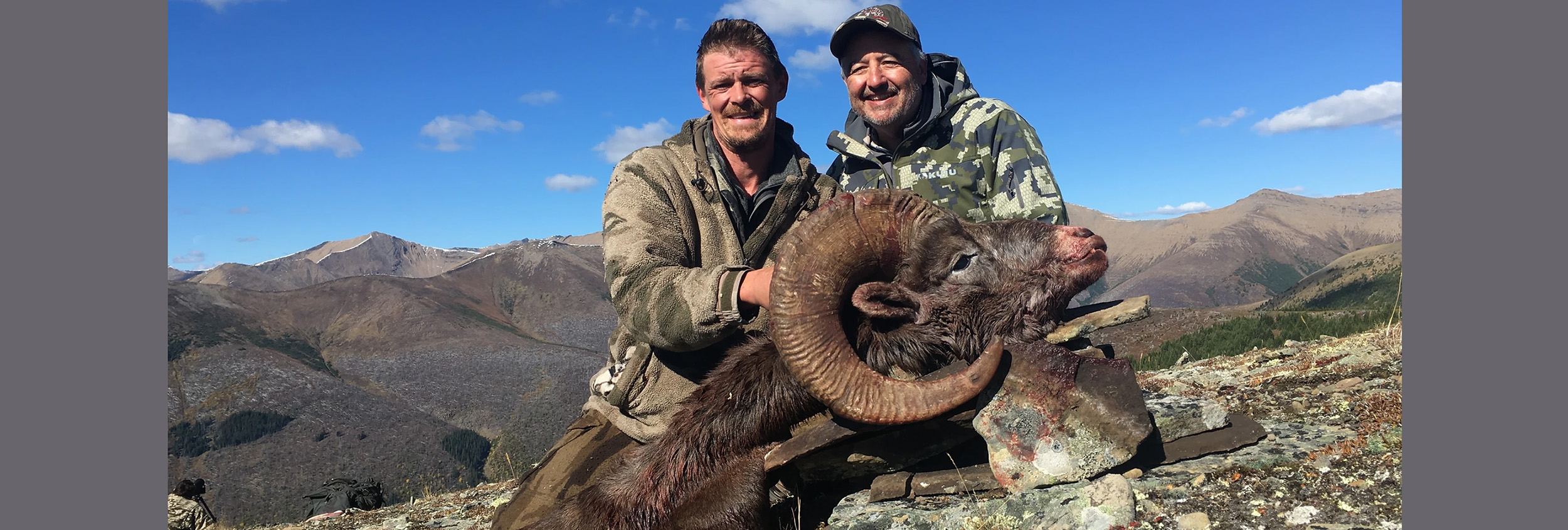 Two hunters pose with a harvested mountain sheep beside rugged alpine mountains under a blue sky.