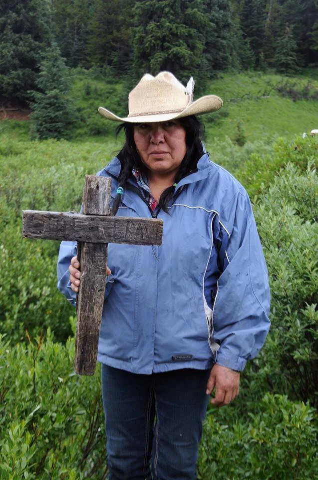 Woman wearing a cowboy hat and blue jacket holding a rustic wooden cross in a forested meadow