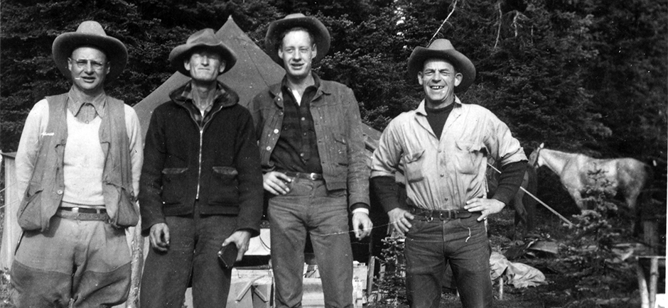 Black-and-white photo of four men in cowboy hats standing outdoors at a forest campsite with tents and a packhorse.