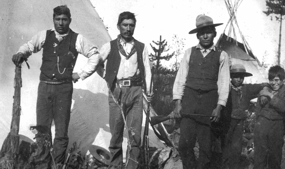 Black-and-white historical photograph of five men standing beside a teepee, with rifles and outdoor gear.
