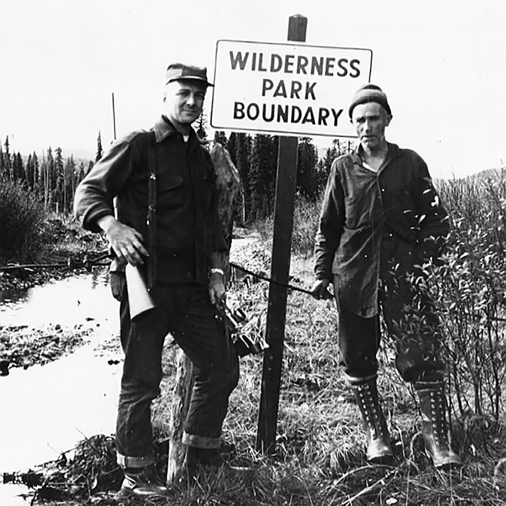 Black-and-white historical photo of two men standing beside a sign reading “Wilderness Park Boundary” in a forested landscape.