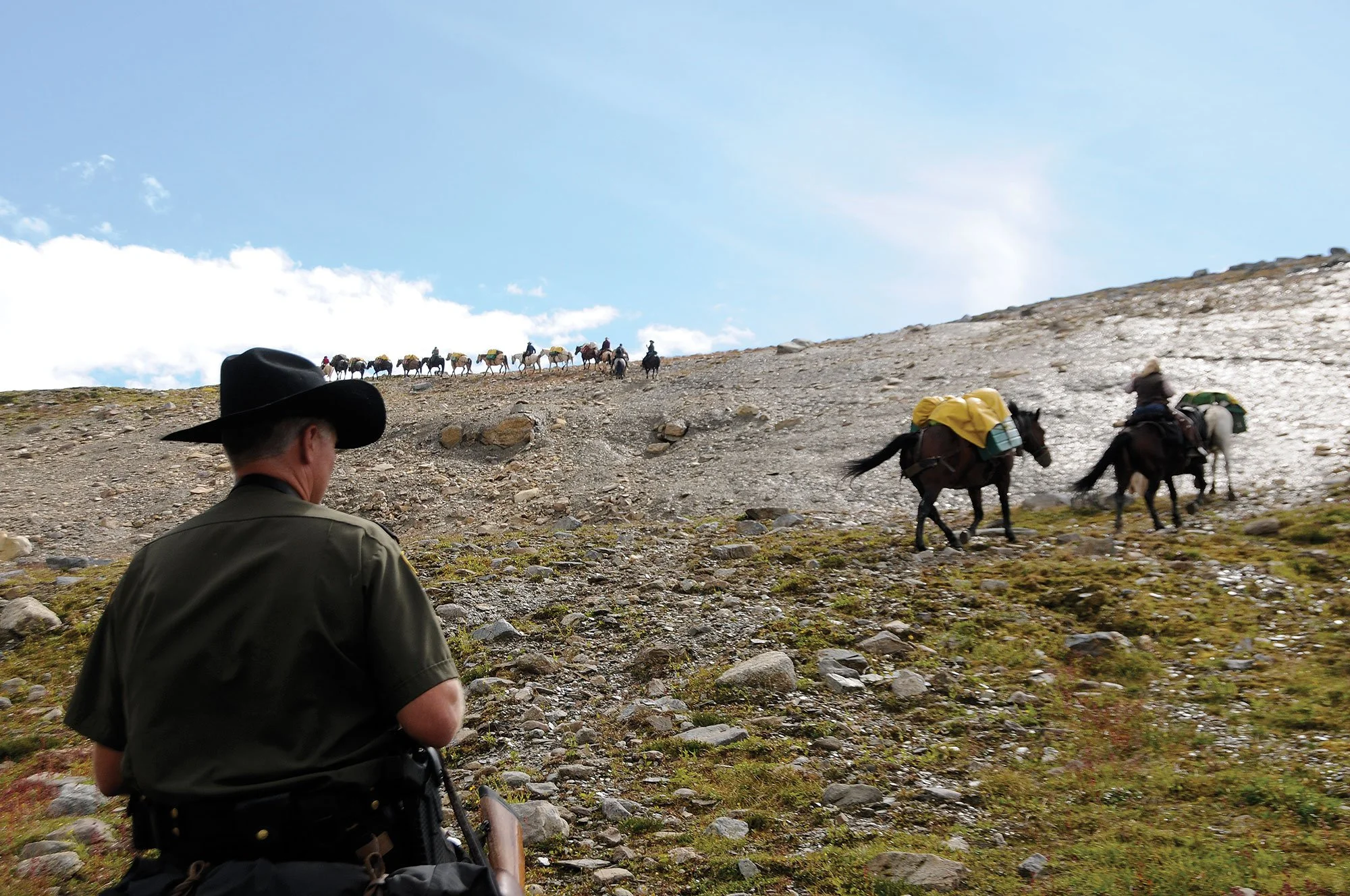 Pack horses and riders crossing a rocky mountain slope beneath a blue sky, with a cowboy in the foreground.