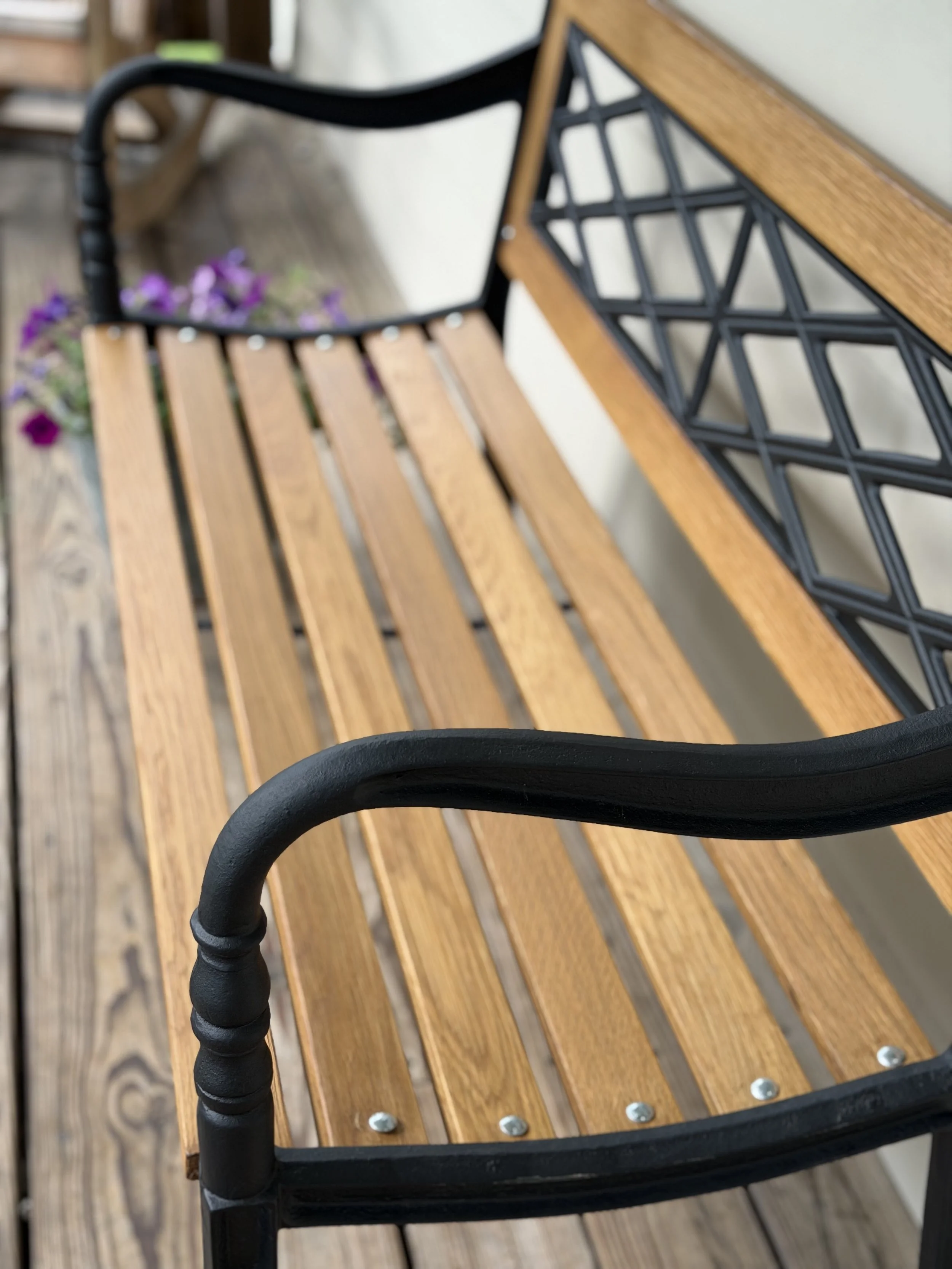 Close-up view of a wooden outdoor bench with black metal armrest and backrest featuring a geometric pattern, placed on a wooden deck with purple flowers in the background.