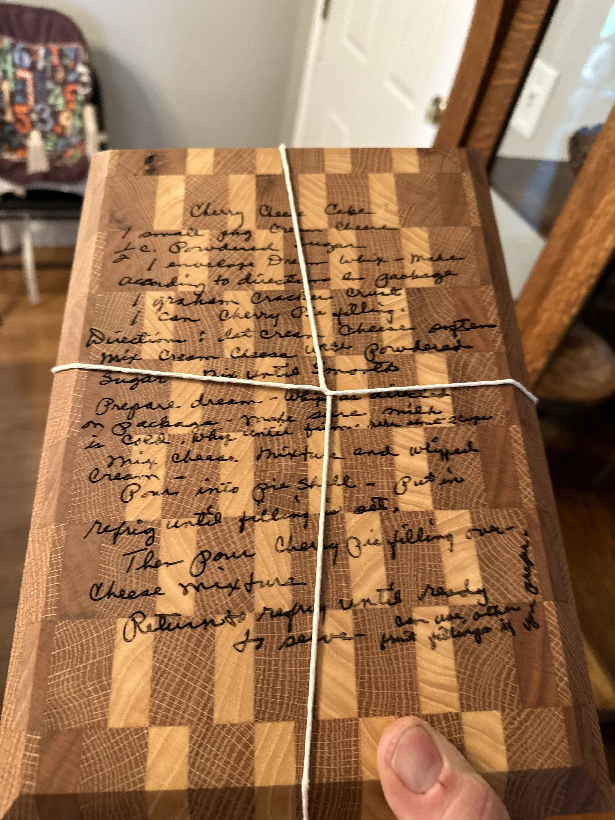 Close-up of a wooden cutting board with recipe instructions handwritten in black ink, held by a person's hand, with a blurred background of a kitchen and a colorful high chair.