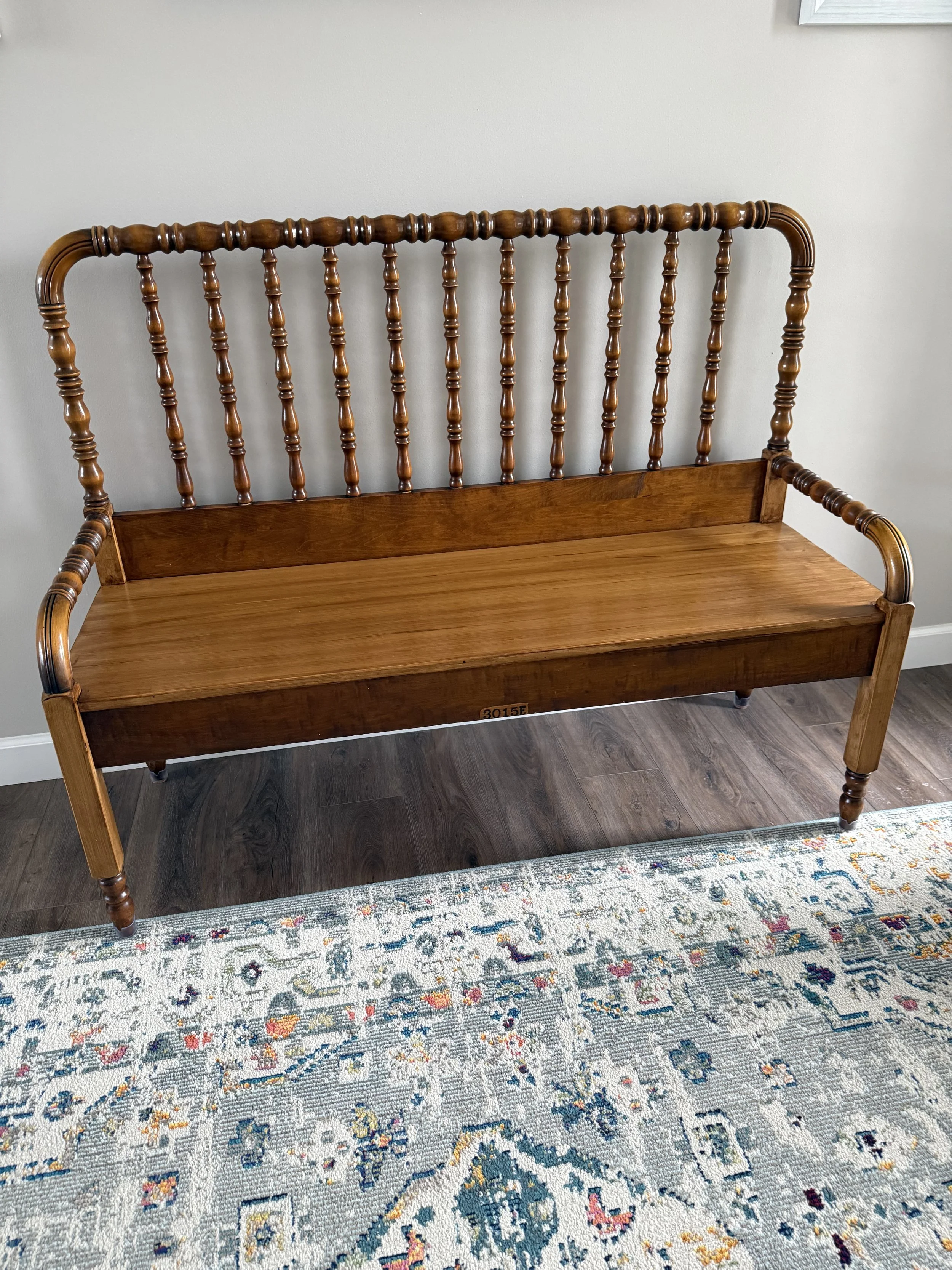 Wooden bench with a tall backrest and spindled design, placed on a multicolored patterned area rug on hardwood flooring.
