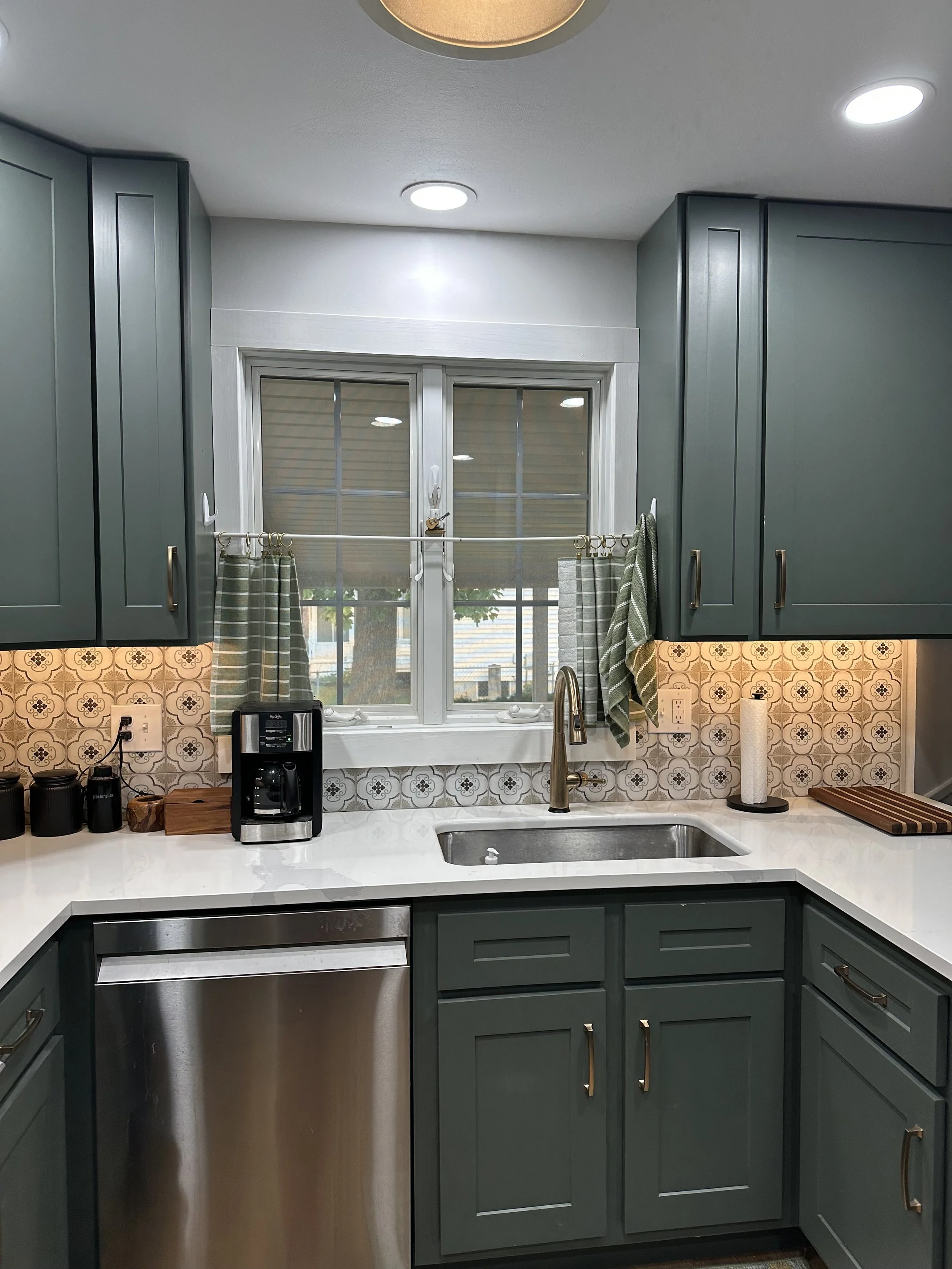 Kitchen with tiled backsplash, gray cabinets, a window above the sink, coffee maker, paper towel holder, and various utensils and containers on the countertop.