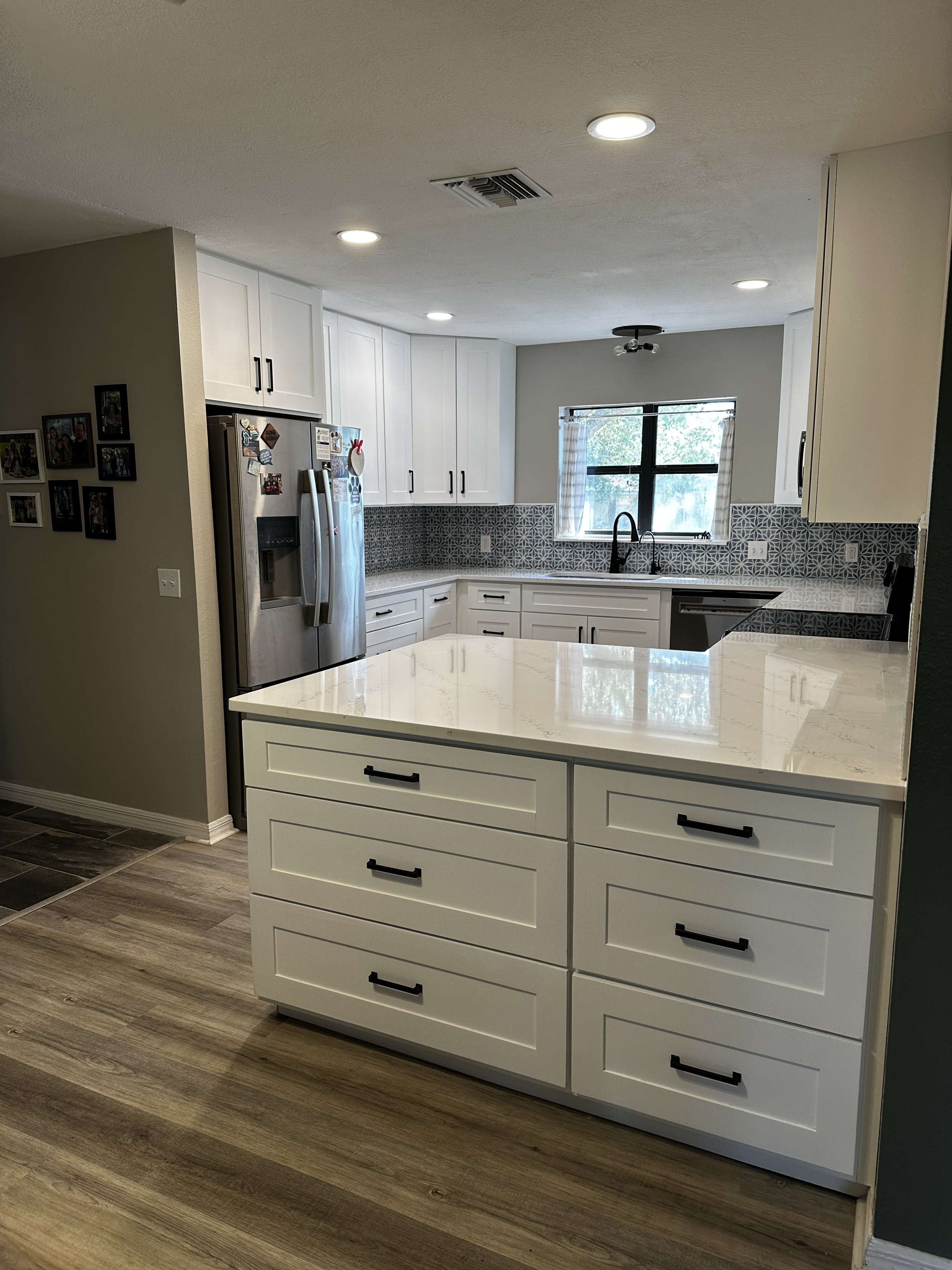 Modern kitchen with white cabinets, black handles, stainless steel refrigerator, and patterned blue and white backsplash, with a large window and wood flooring.
