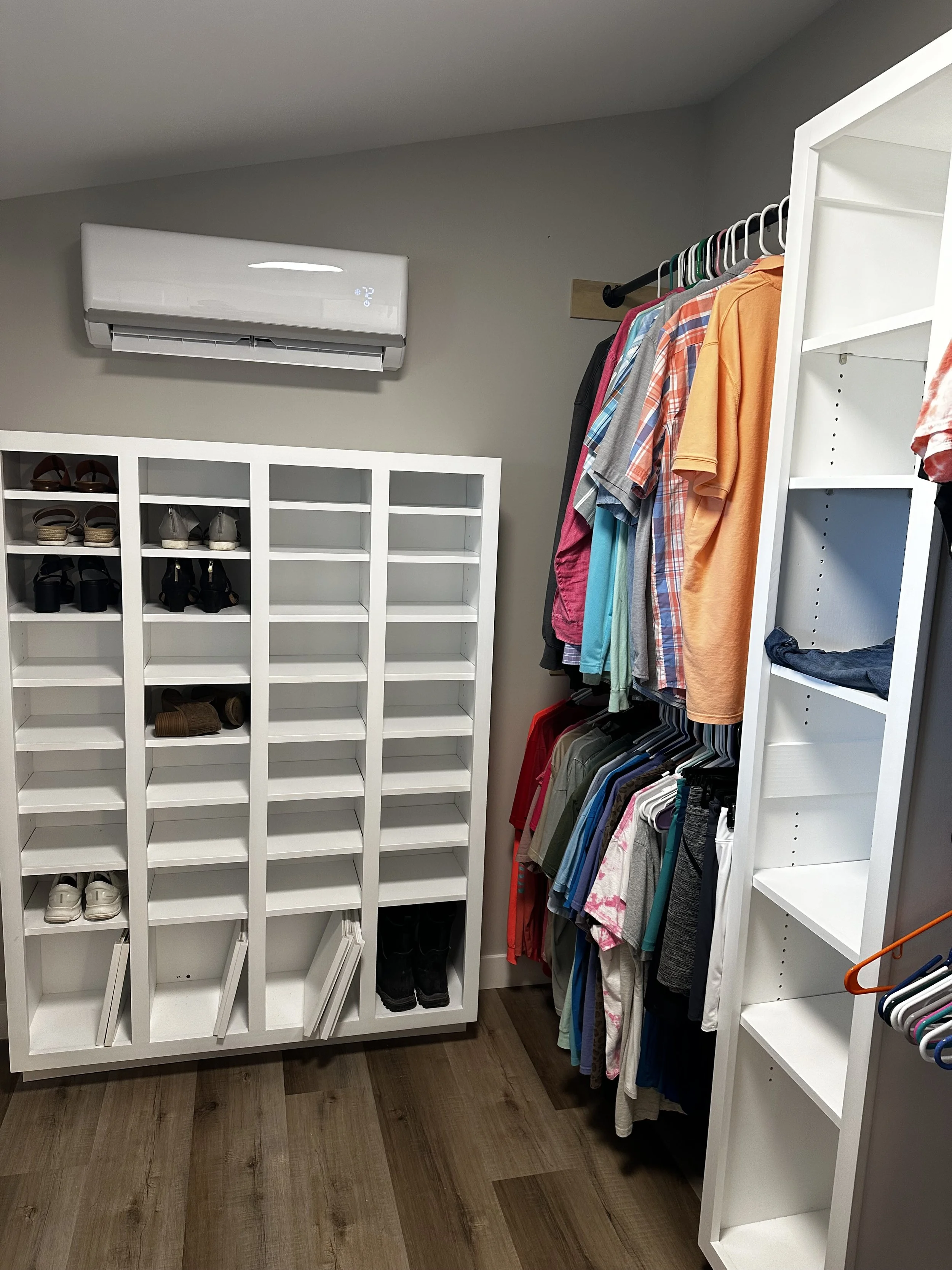 A walk-in closet with white shelves and a clothing rack holding various shirts. Shoes are stored on the shelves and the floor.