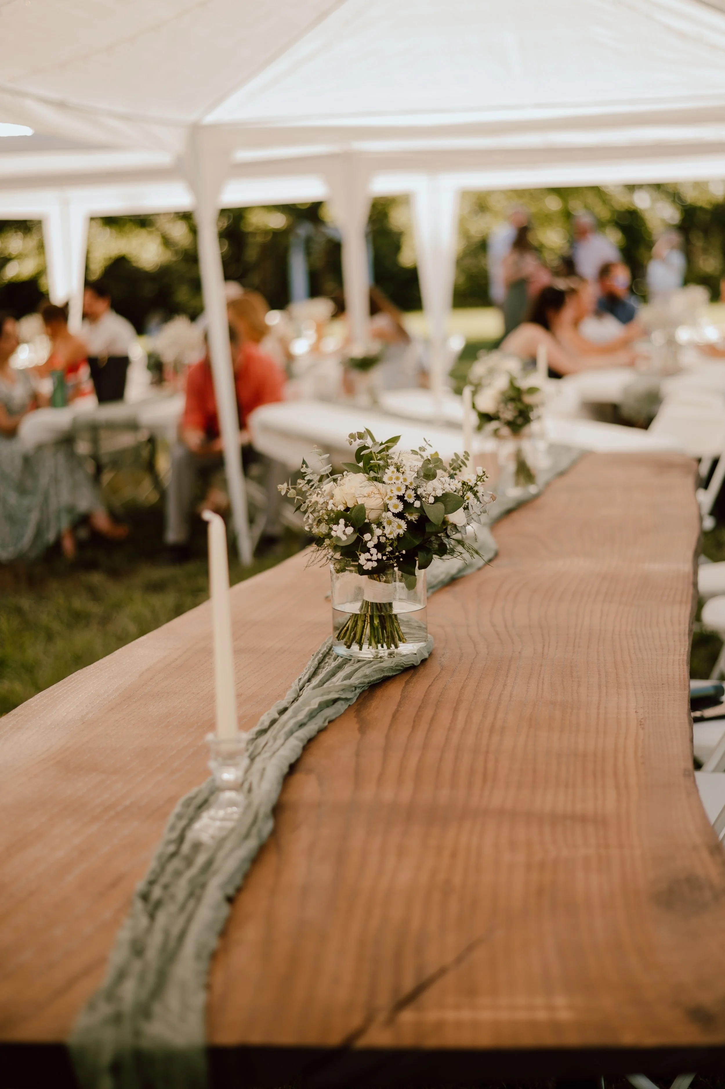 Close-up of a wooden table with a cloth runner and small flower arrangements, set for an outdoor event under a white canopy with people seated in the background.