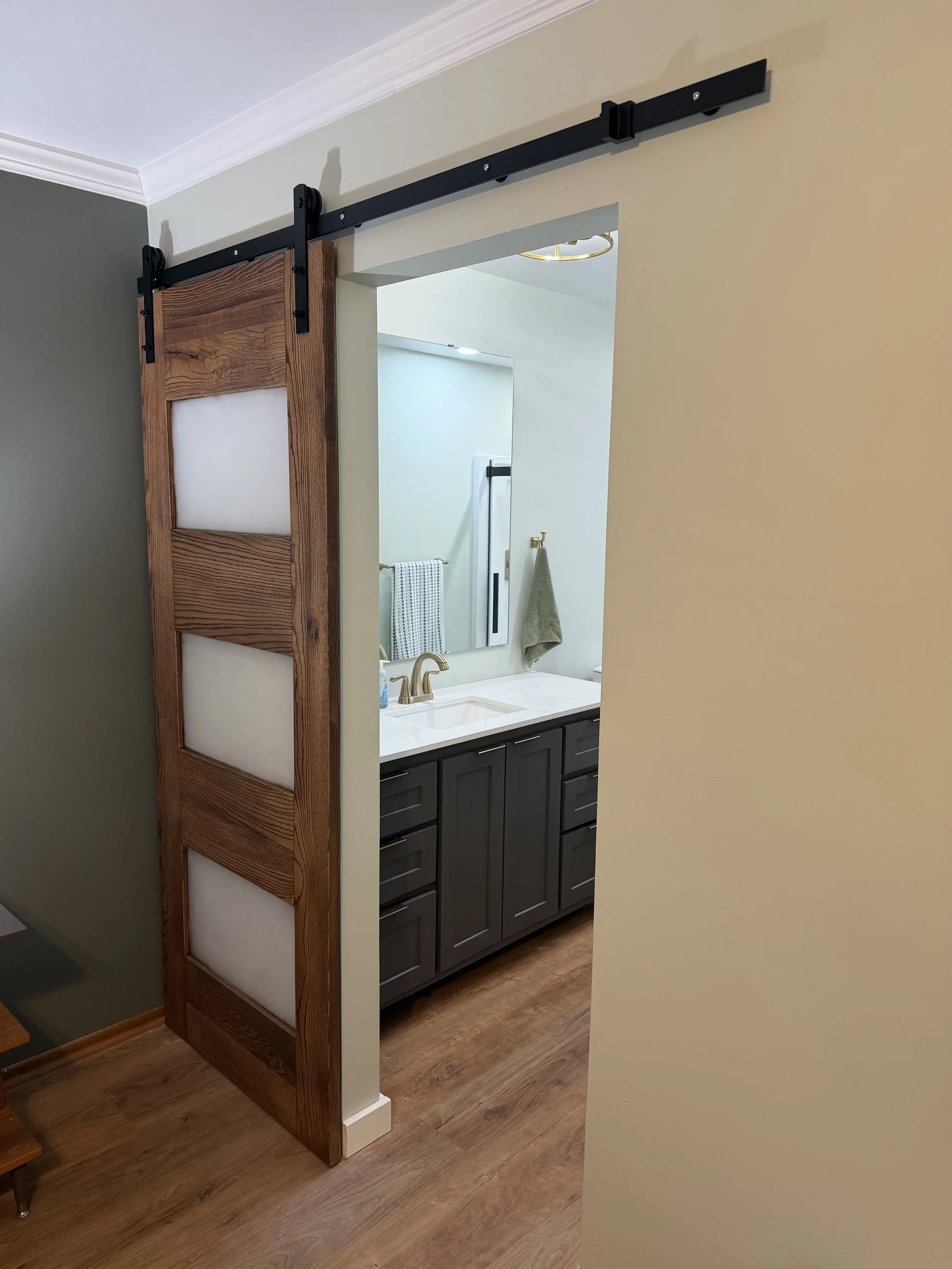 View through a doorway revealing a bathroom with a white countertop, a gold faucet, dark gray cabinets, a mirror, a towel, and a towel bar visible in reflection.