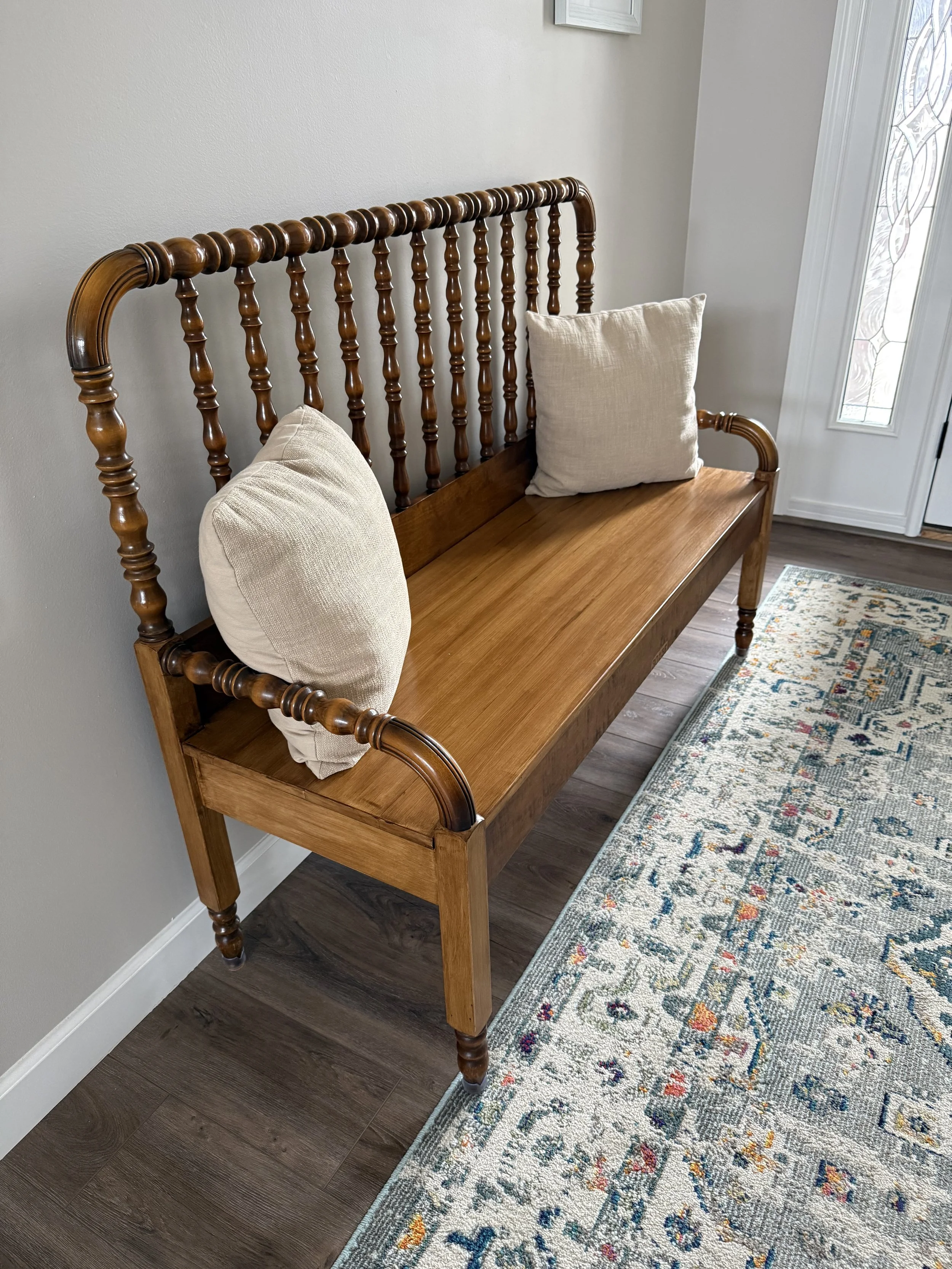 A wooden bench with turned spindles and armrests, two beige cushions placed on it, positioned next to a door with a decorative glass window. A patterned rug is partially visible on the floor beneath the bench.