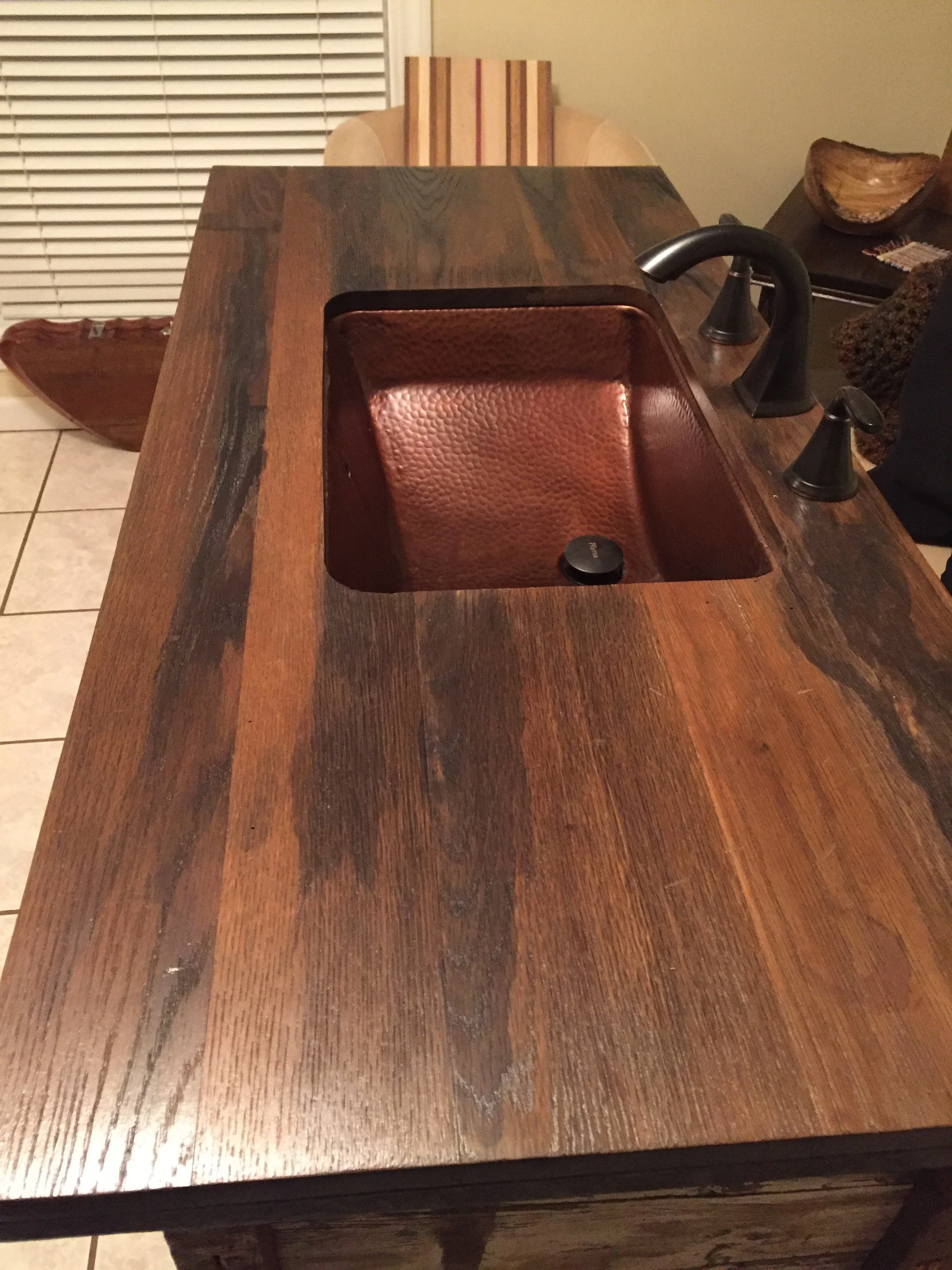 Close-up of a wooden kitchen or dining table with a built-in sink, a copper-colored chair in the background, and a side table with decorative bowls.