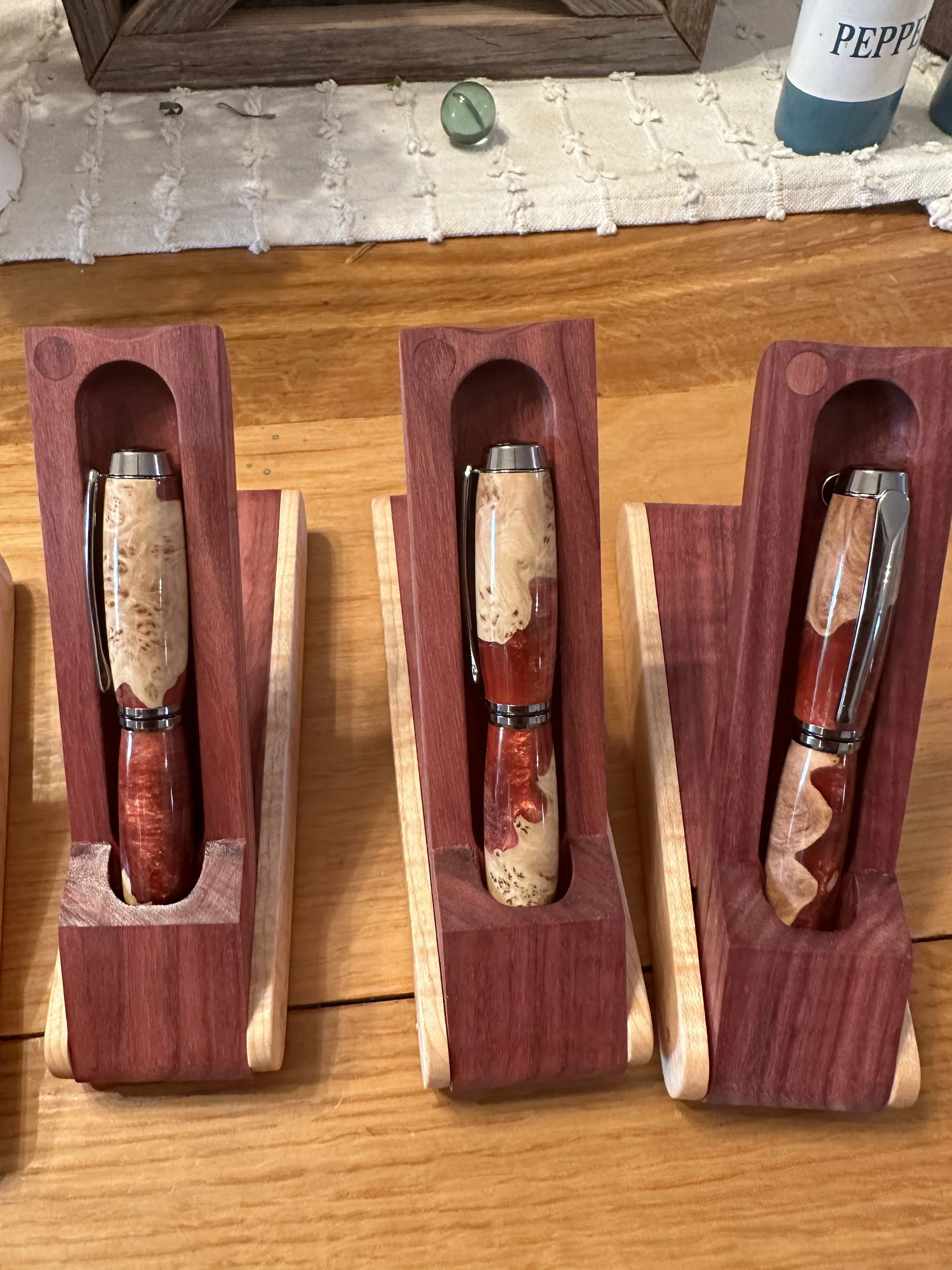 Three handmade pens with wood and resin bodies, resting in wooden display holders on a wooden table.