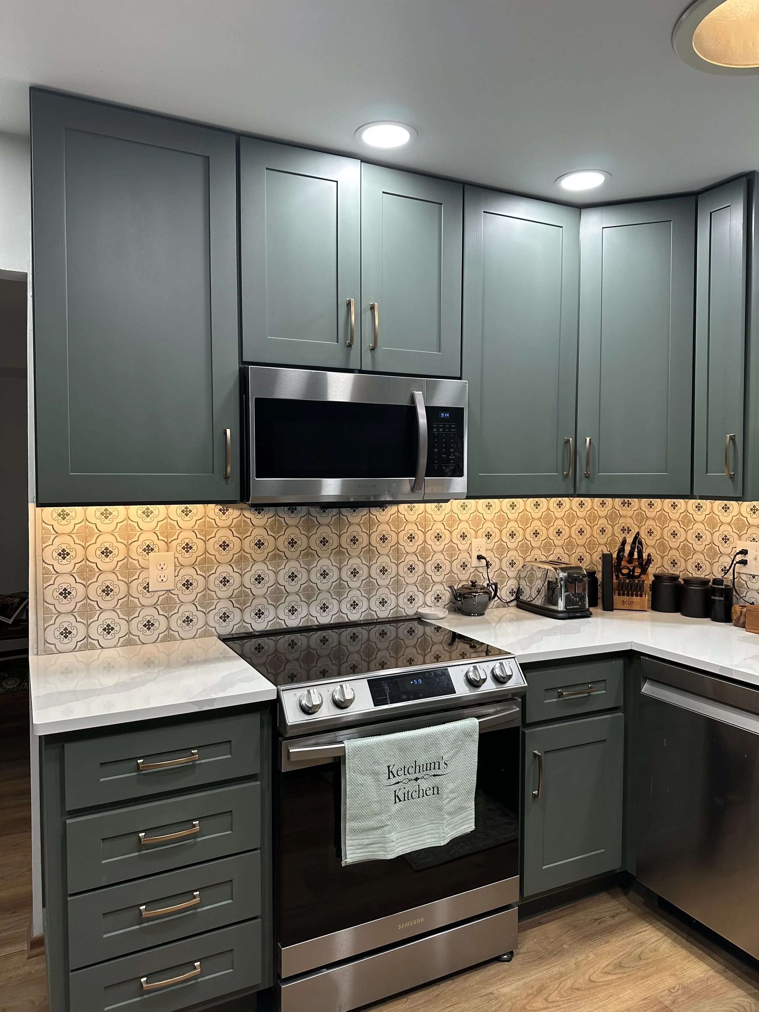 A kitchen with green cabinets, a stainless steel microwave, and a stove with a beige and brown patterned tile backsplash. There is a dish towel hanging from the oven handle that reads "Ketchum's Kitchen." Small kitchen appliances and utensils are on 