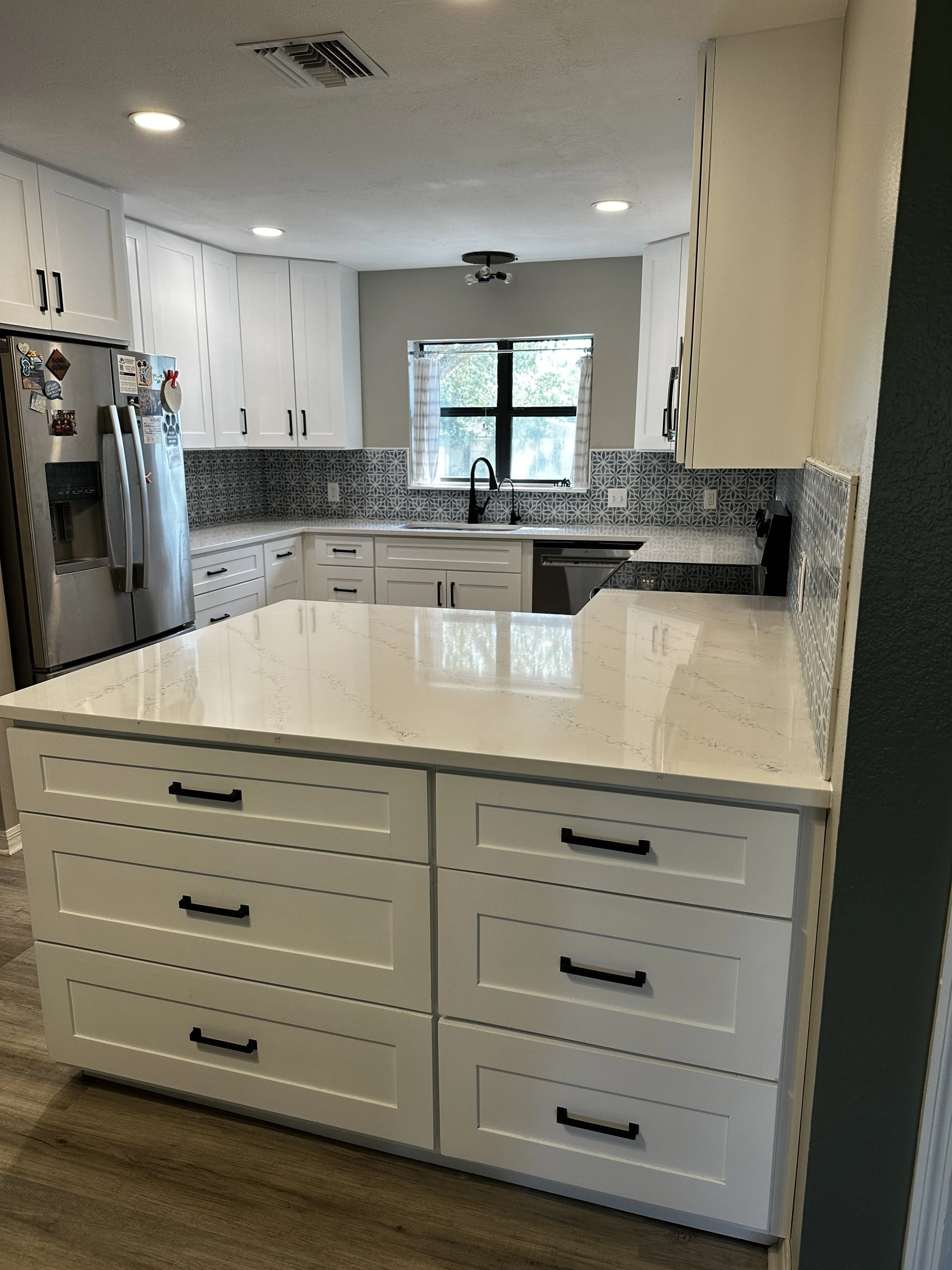 Modern kitchen with white cabinets, black hardware, a countertop island, and a patterned backsplash in gray and white. There's a window with checkered curtains, a stainless steel refrigerator with magnets, and a black stove. Recessed ceiling lights a