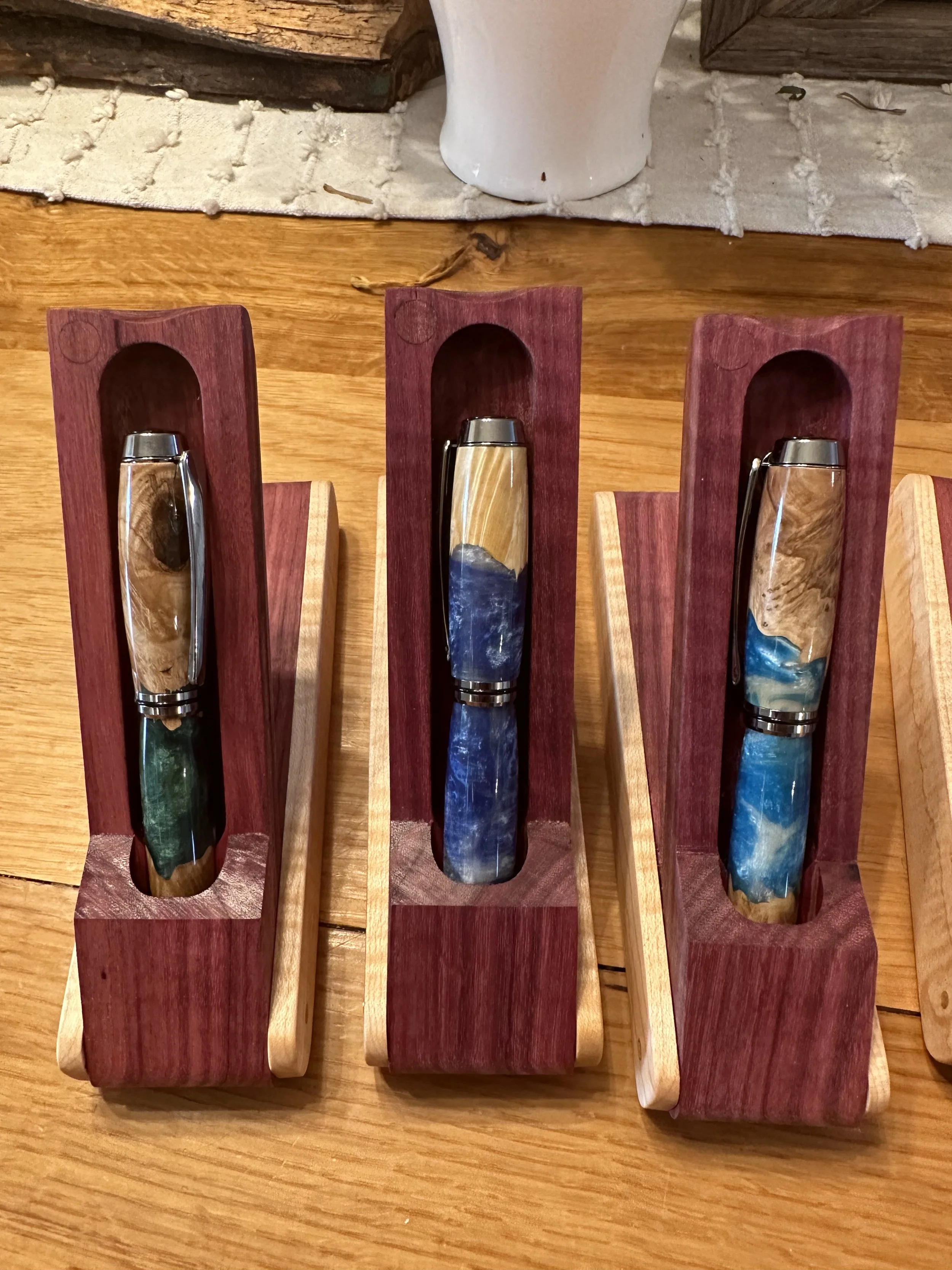 A display of five handcrafted pens in custom wooden holders, with varying wood grains and colors, placed on a wooden table.