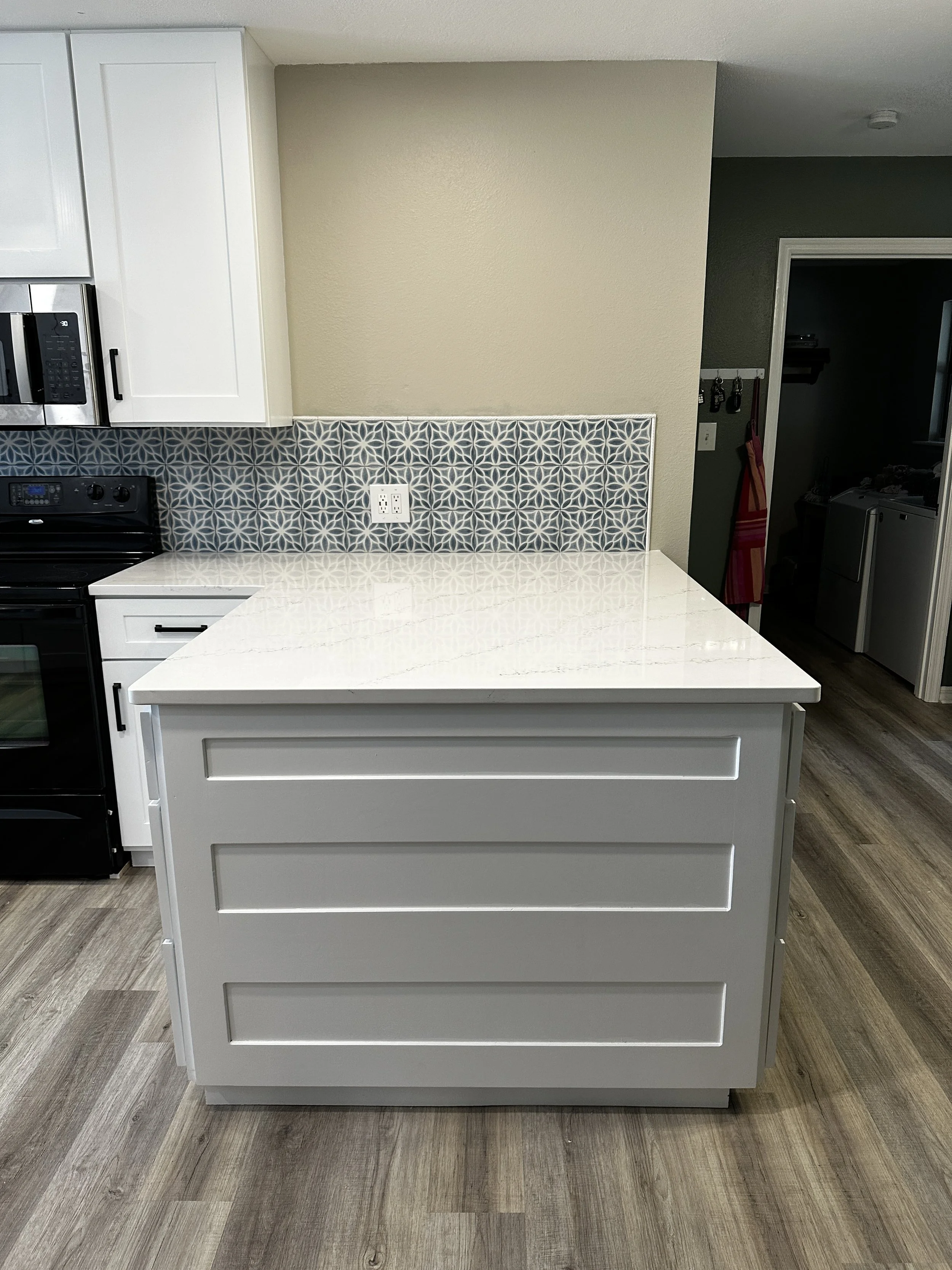 White kitchen island with drawers, part of a kitchen with patterned backsplash, adjacent to a stove and microwave, and a wall outlet visible on the backsplash.
