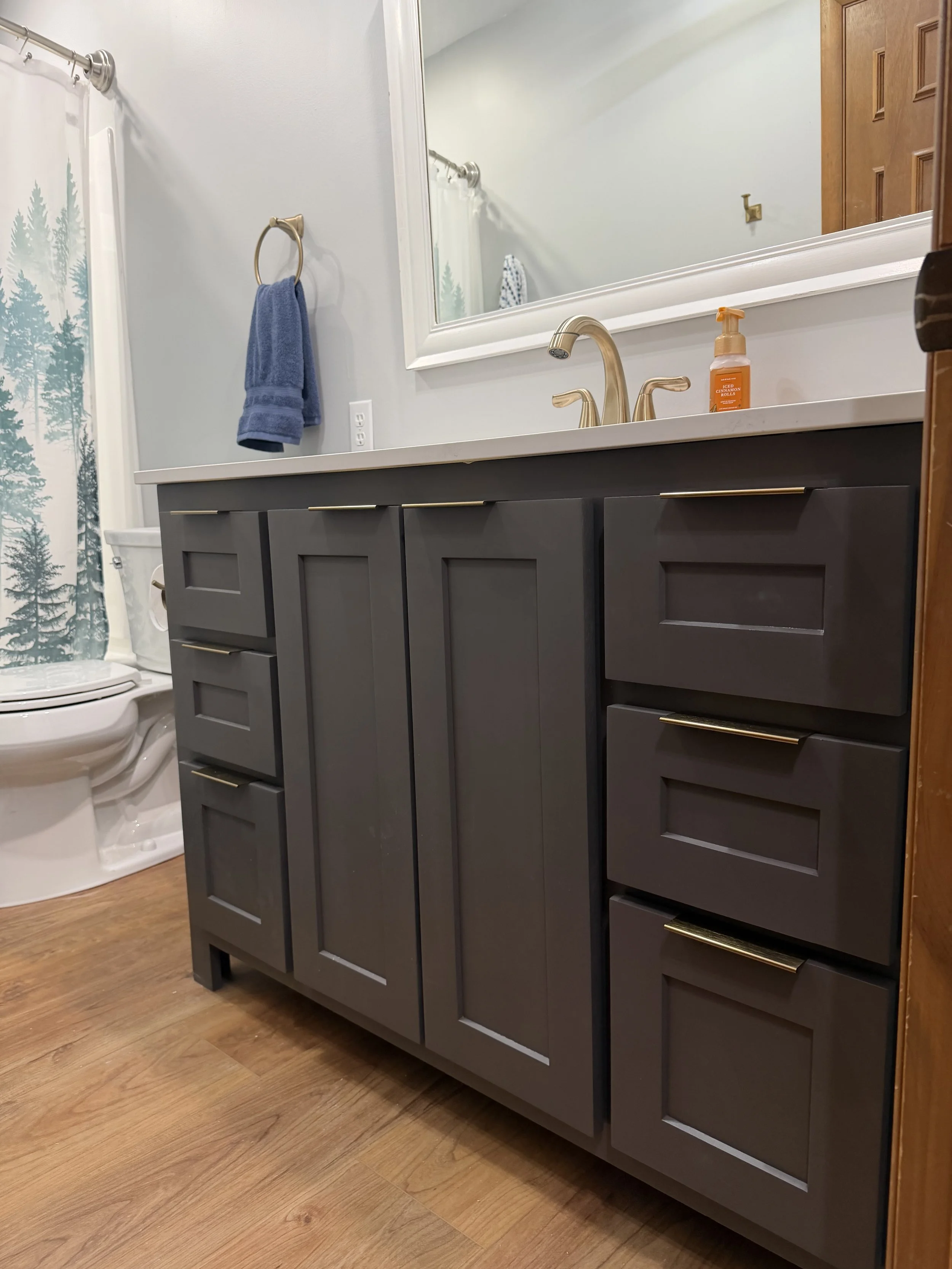 A bathroom vanity with a dark gray cabinet, gold handles, a white countertop with a gold faucet, a soap dispenser, a large mirror, and a towel ring with a blue towel. Behind the vanity, there is a partial view of a toilet with a decorated shower curtain and a wooden floor.