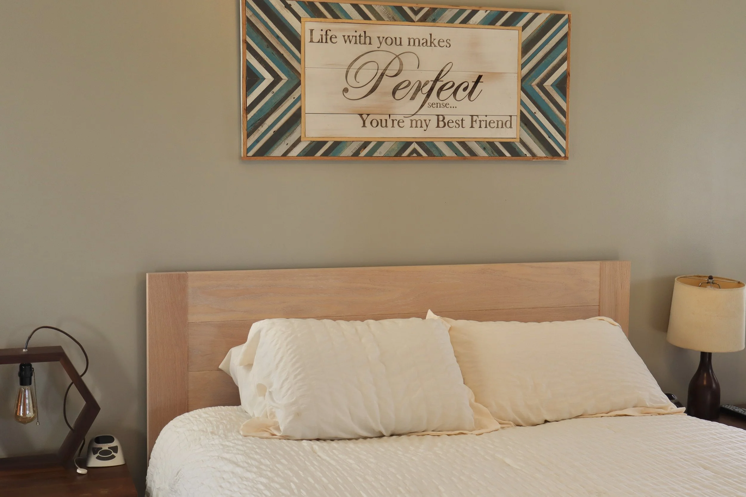 A bedroom with a wooden headboard, a side table with a lamp, and a framed wall art sign that reads 'Life with you makes perfect sense... You're my best friend.' The bed has white quilted bedding with pillows. A handheld device and a small speaker are