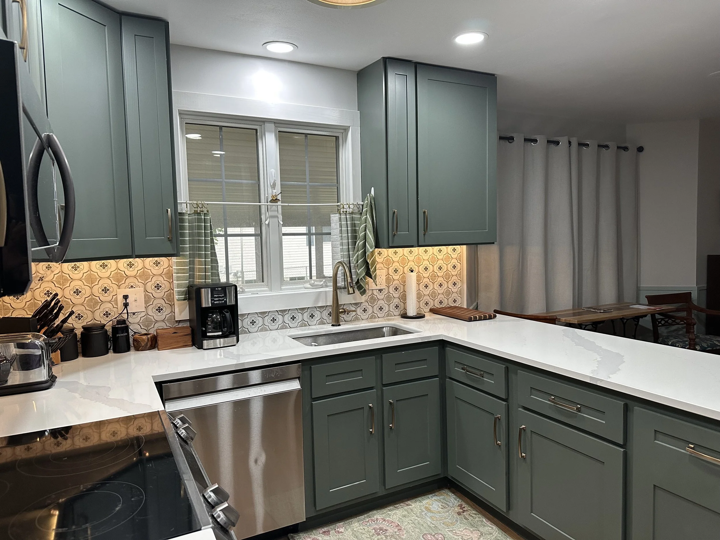 Kitchen with green cabinets, white countertop, window above sink with curtains, patterned backsplash, and various kitchen appliances and utensils.