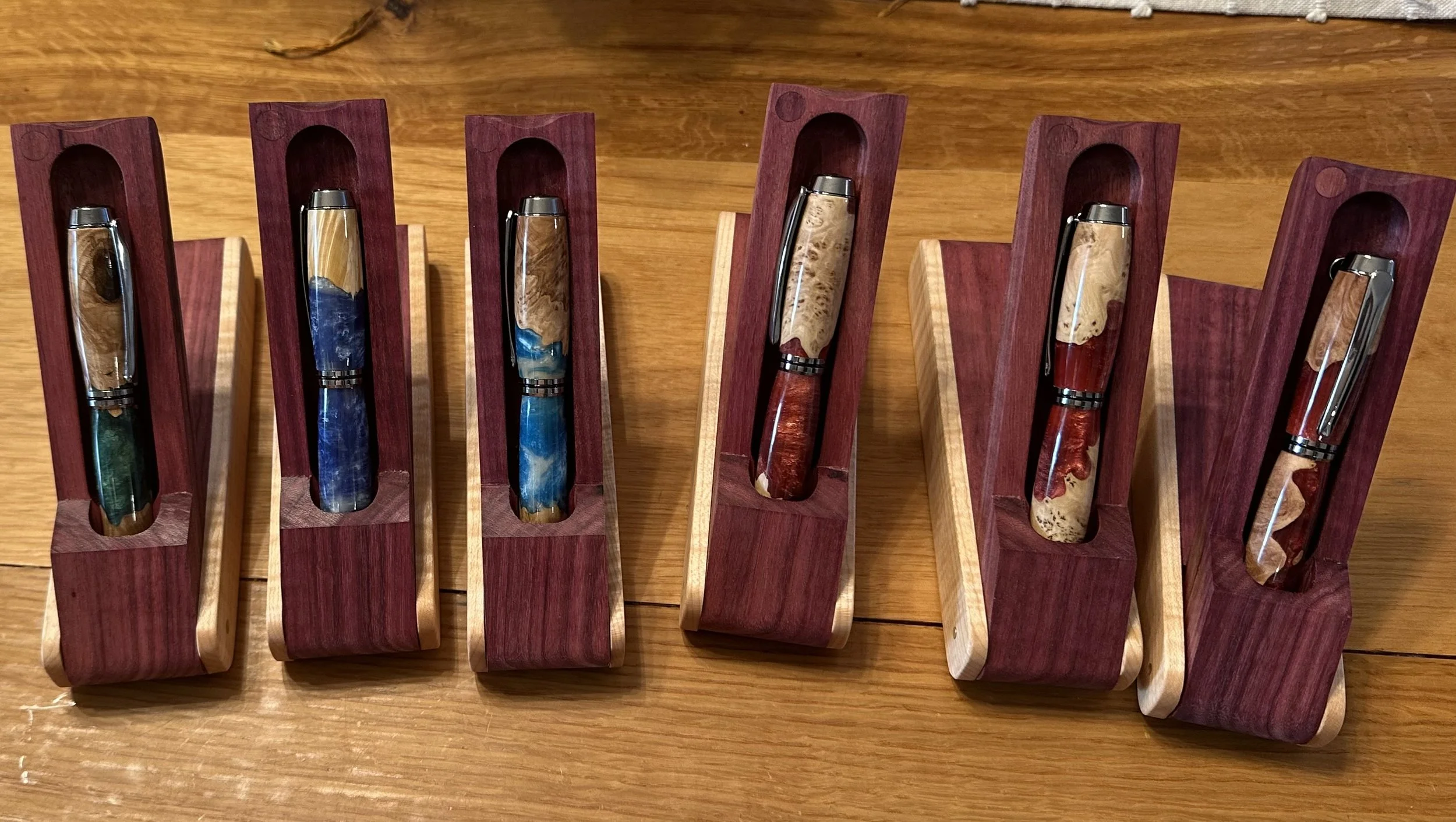 Six handcrafted pens displayed in individual wooden cases on a wooden surface.