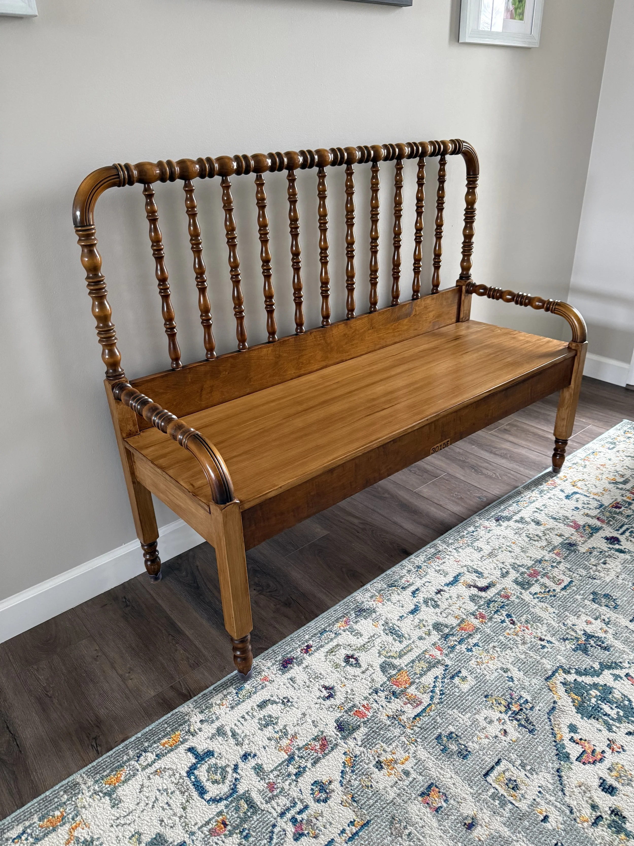 A wooden bench with a high backrest and armrests, placed on a hardwood floor next to a patterned rug in a room with light-colored walls and framed pictures on the wall.
