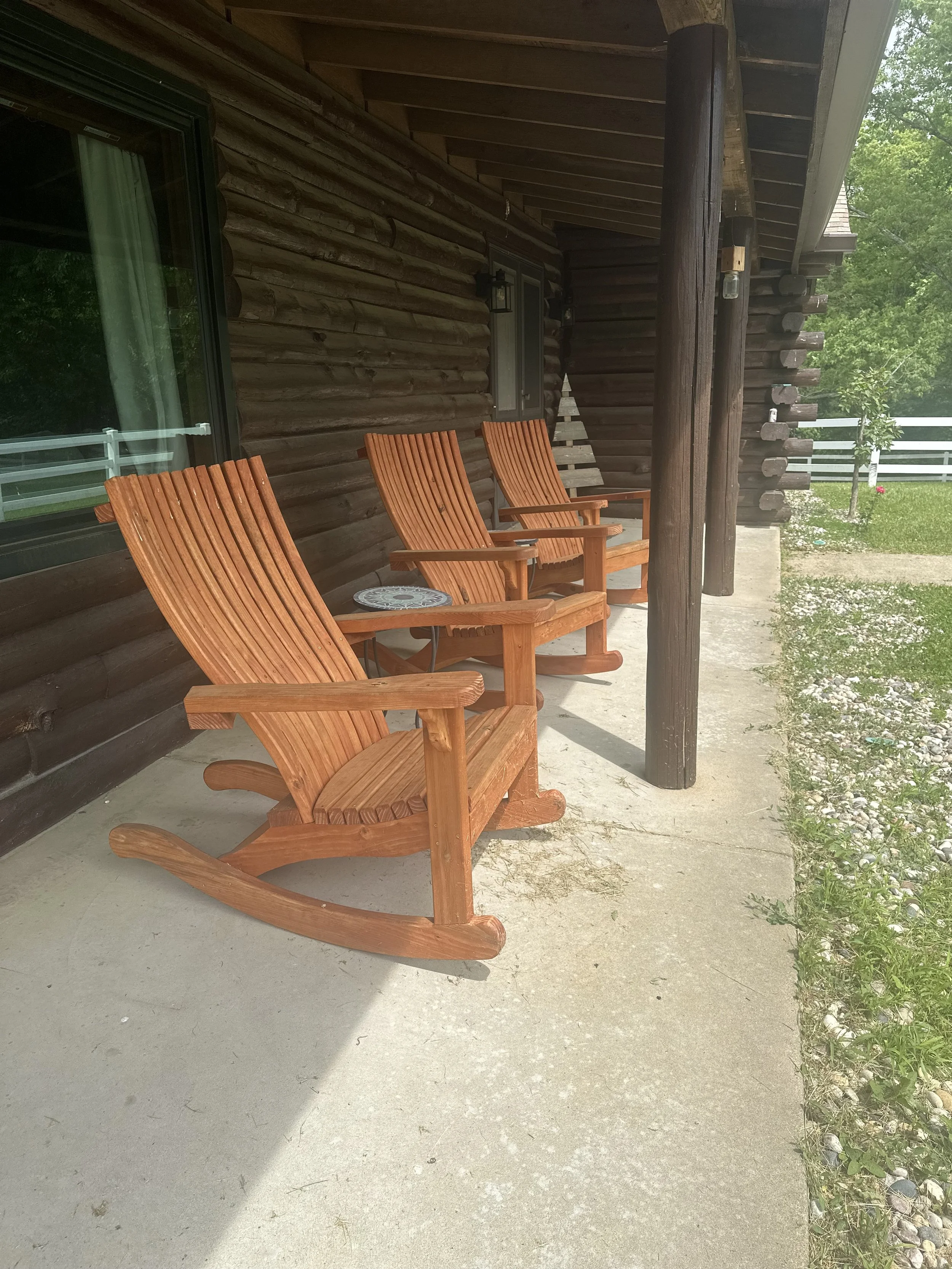 Three wooden rocking chairs on a porch next to a wooden house with a large window, surrounded by green trees and grass.