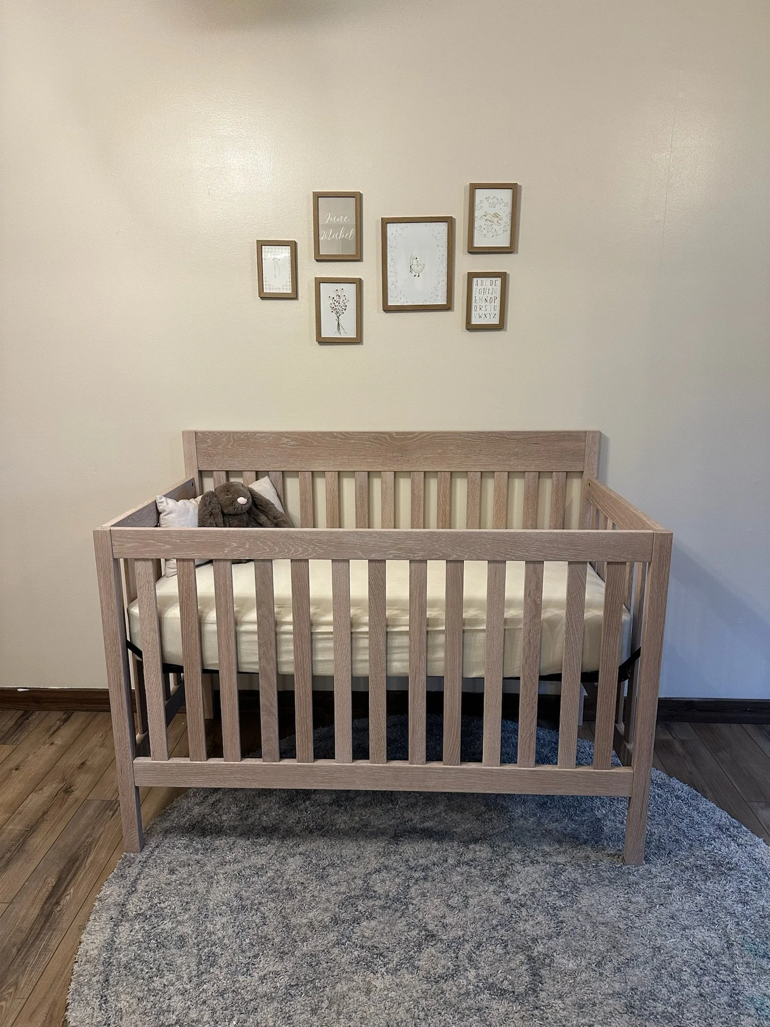 A wooden crib with a stuffed animal and pillows inside, placed on a gray rug in a room with light-colored walls decorated with framed artwork.