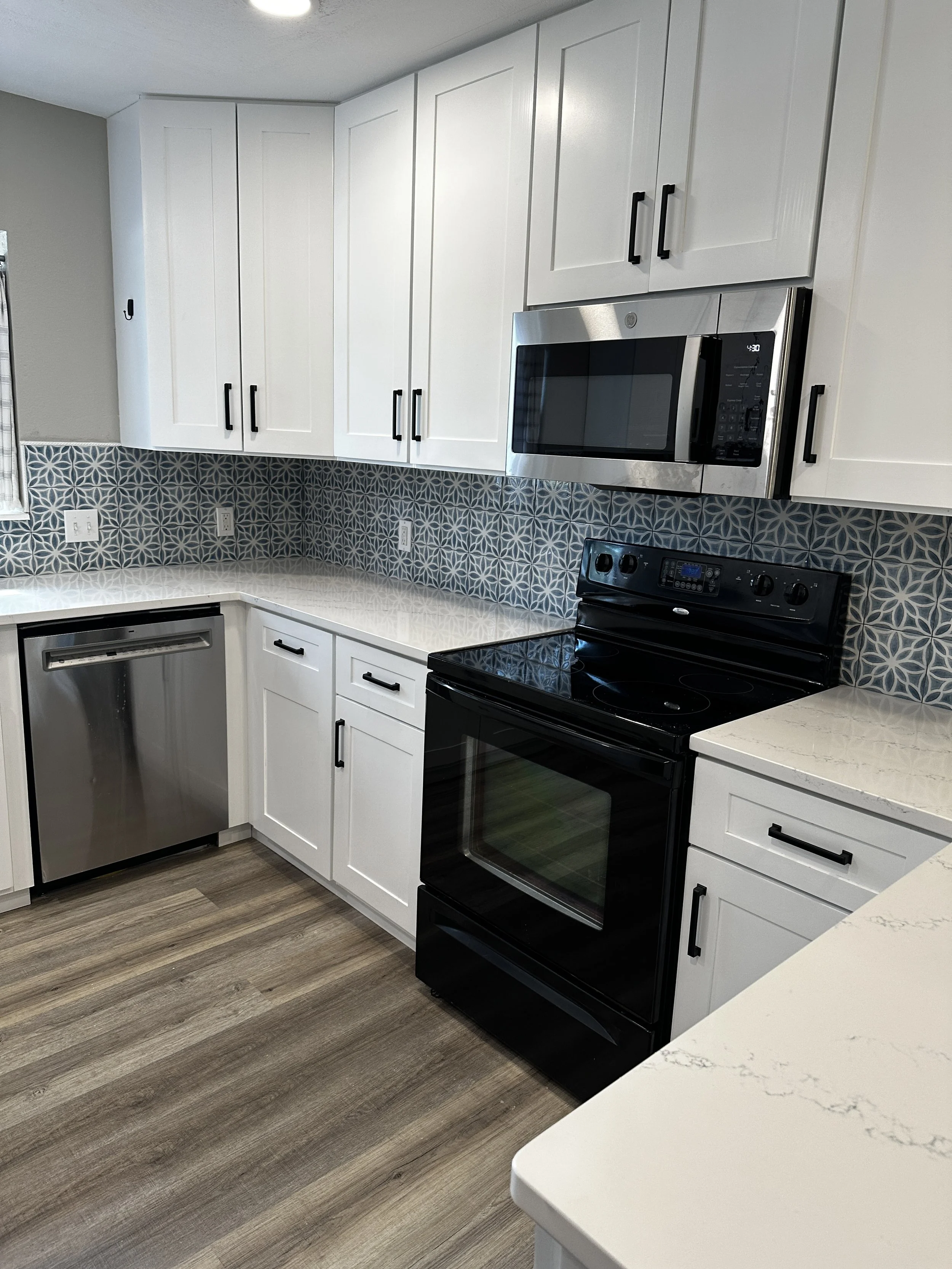 A modern kitchen with white cabinets, black handles, a black electric stove, stainless steel microwave, a dishwasher, wooden flooring, and white marble countertops with gray veining.