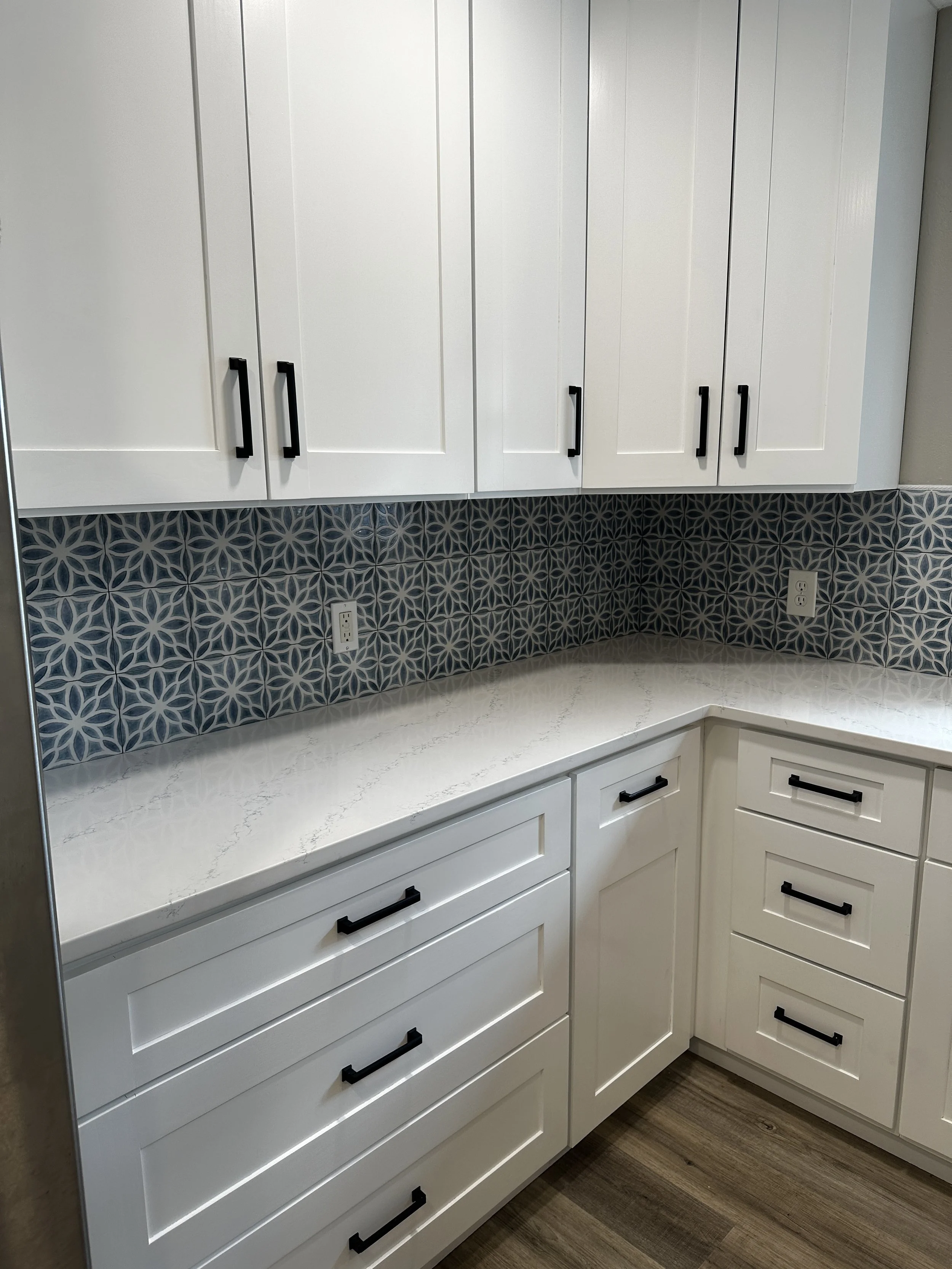 White kitchen cabinets with black handles, patterned tile backsplash, white countertop, and electrical outlets on the wall.