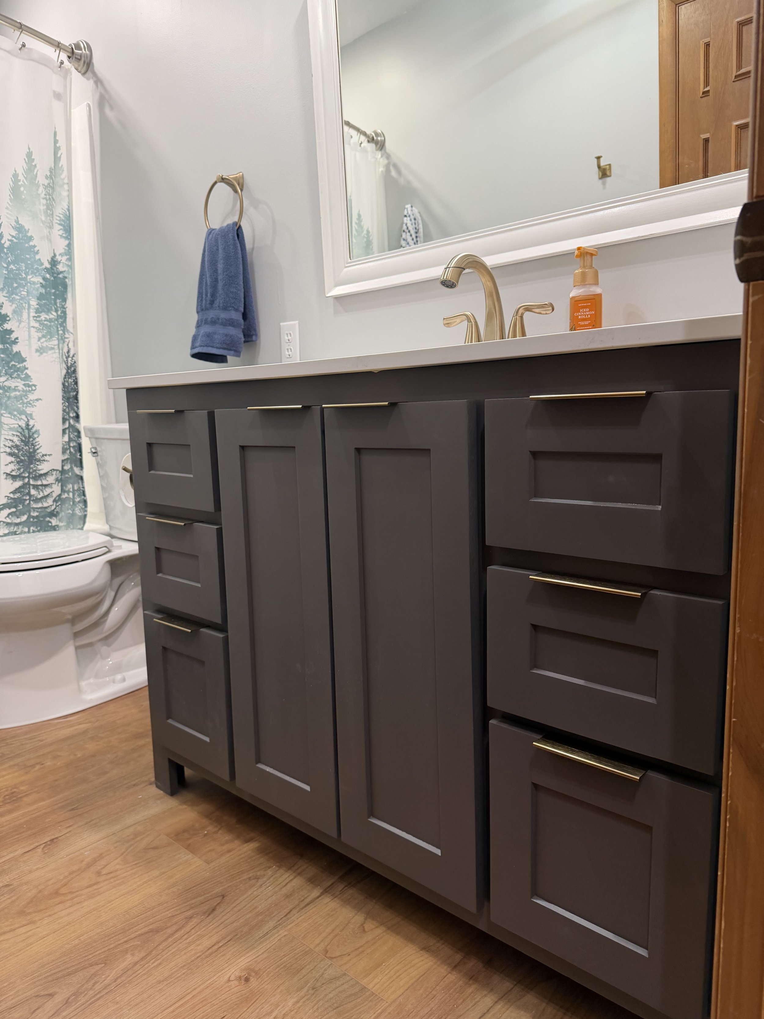 Bathroom with gray vanity, gold handles, a white countertop, large wall mirror, gold faucet, and a bottle of soap or sanitizer on the counter. In the background, there is a toilet and shower with a tree-patterned curtain.
