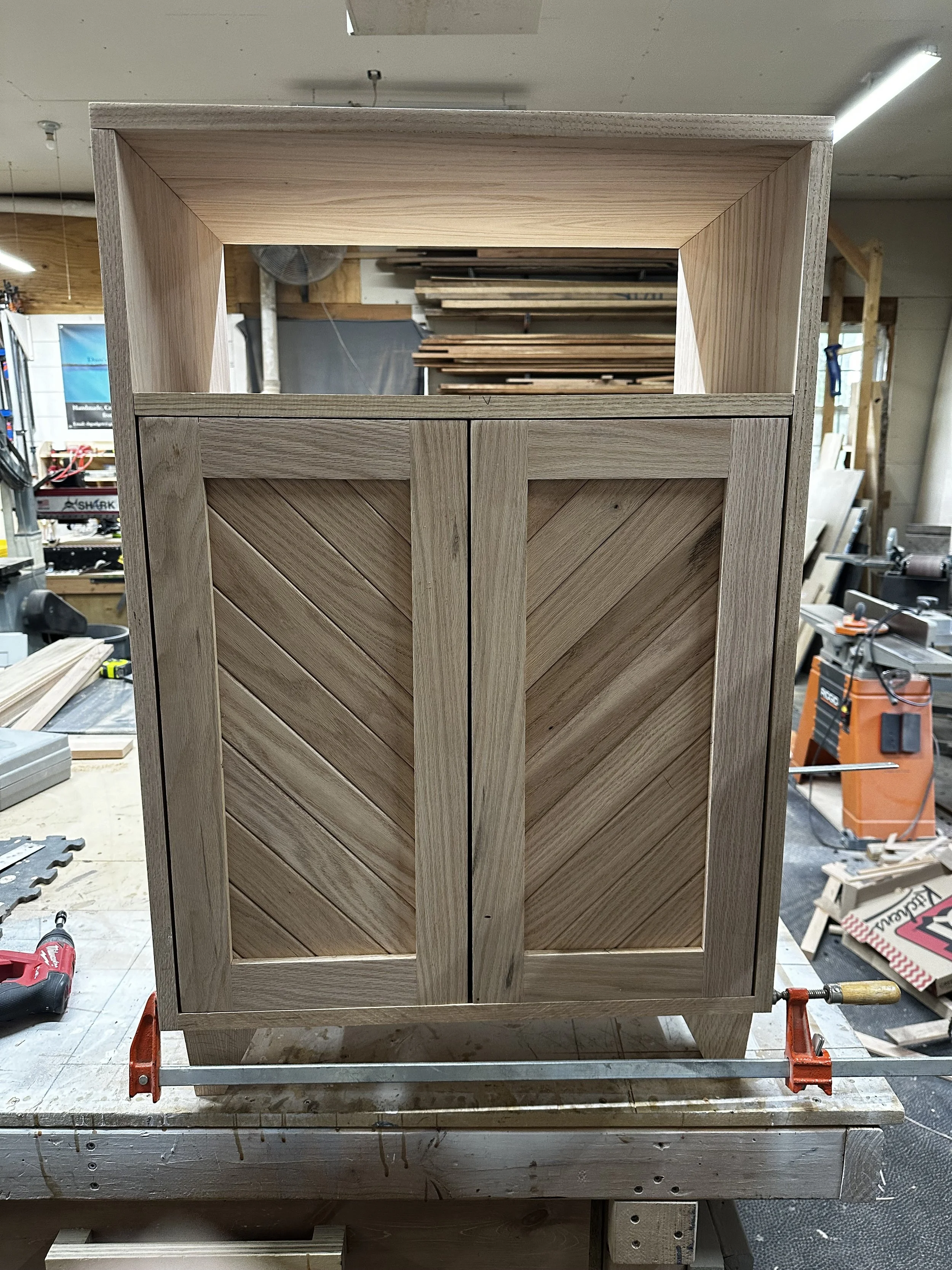 Wooden cabinet with chevron pattern on the doors, in a woodworking shop, partially assembled, with various tools and wood pieces around.
