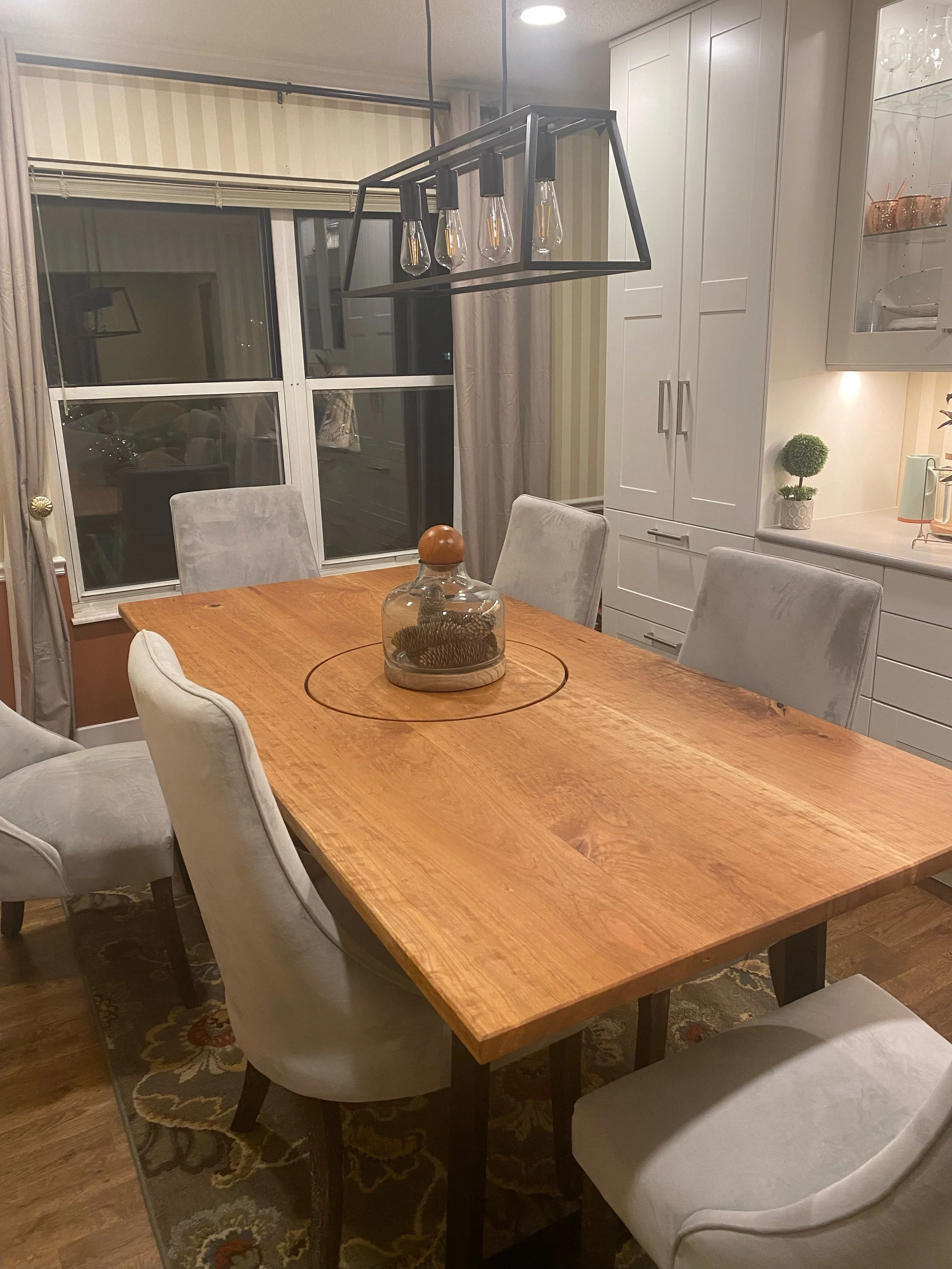 A dining room with a wooden table surrounded by white upholstered chairs. In the background, there are white cabinets and a glass display with glassware. A modern black chandelier with exposed bulbs hangs above the table. A glass jar with pinecones is on the table, and a decorative rug is underneath.