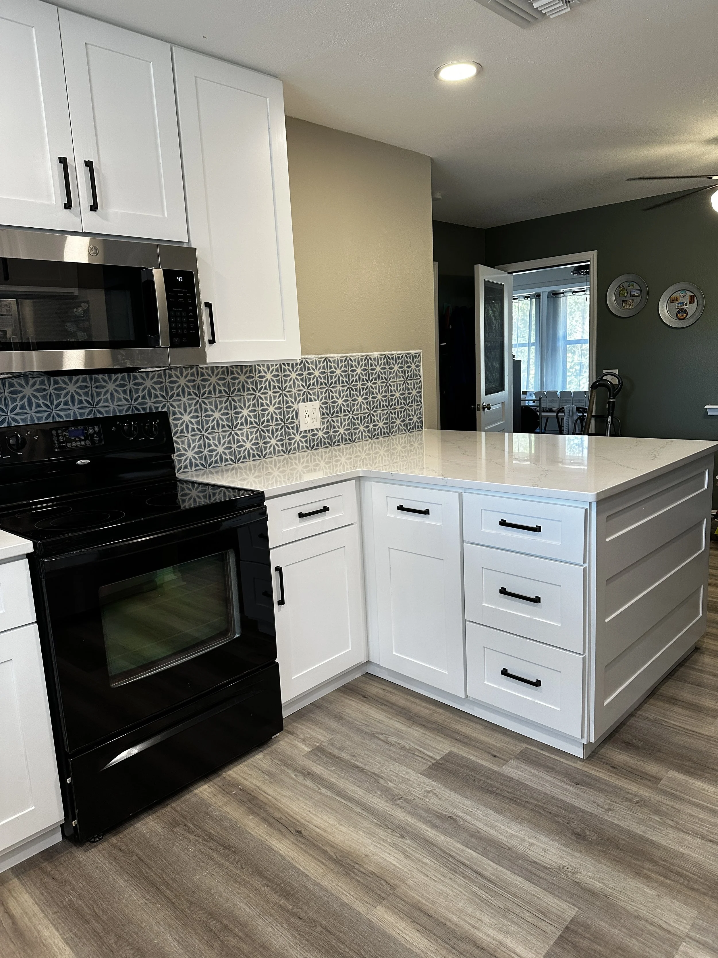 Modern kitchen with white cabinets, a black stove, a stainless steel microwave, geometric patterned backsplash, and a white island with drawers. In the background, there is a doorway leading to a room with a large window and blue curtains.