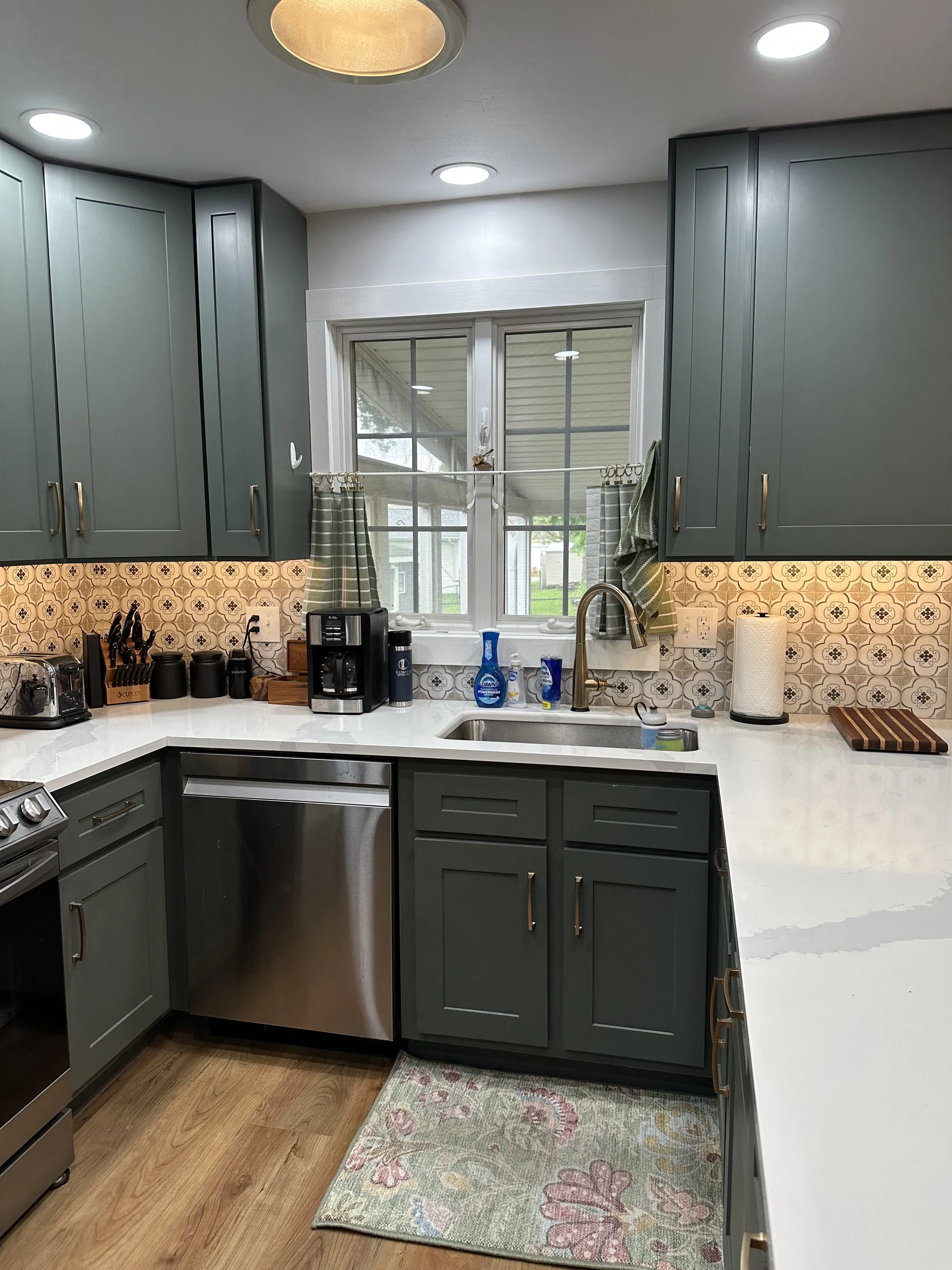 A kitchen with blue cabinets, patterned tile backsplash, white countertops, a window with curtains, and various appliances and utensils on the counter.