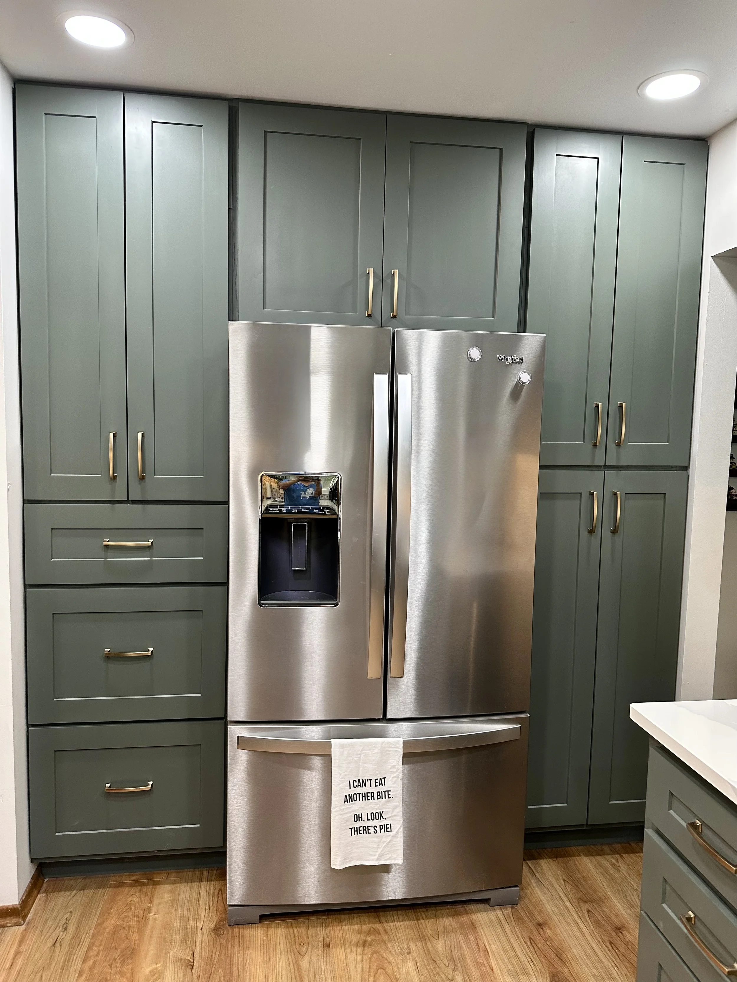 Kitchen with gray cabinets surrounding a stainless steel side-by-side refrigerator with a water and ice dispenser. A cloth with a humorous message hangs on the refrigerator handle. The floor is wooden and there is a white countertop on the right.