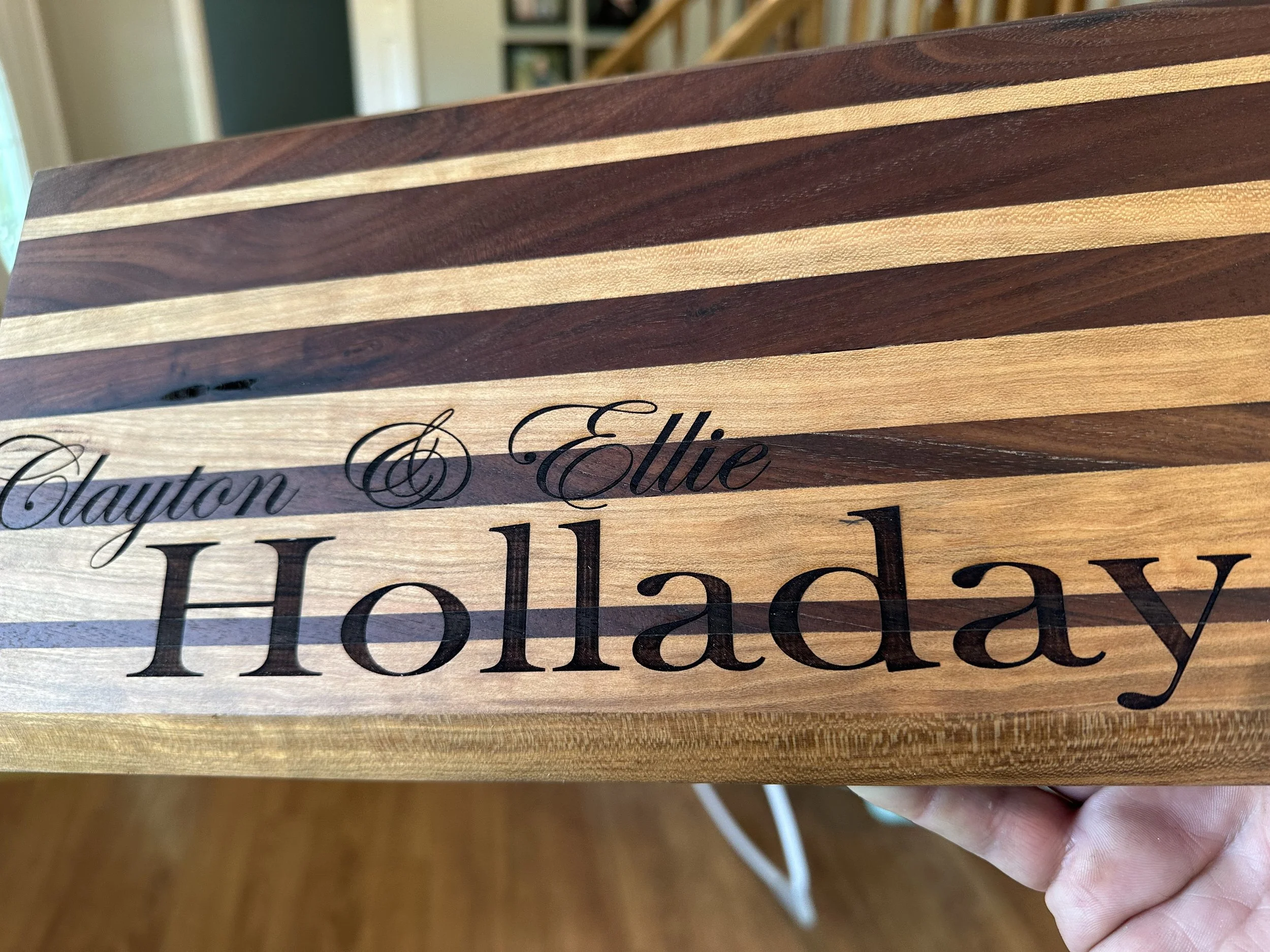 Close-up of a wooden sign with alternating light and dark wood stripes, featuring engraved black cursive text reading 'Elie' and large capitalized letters below.