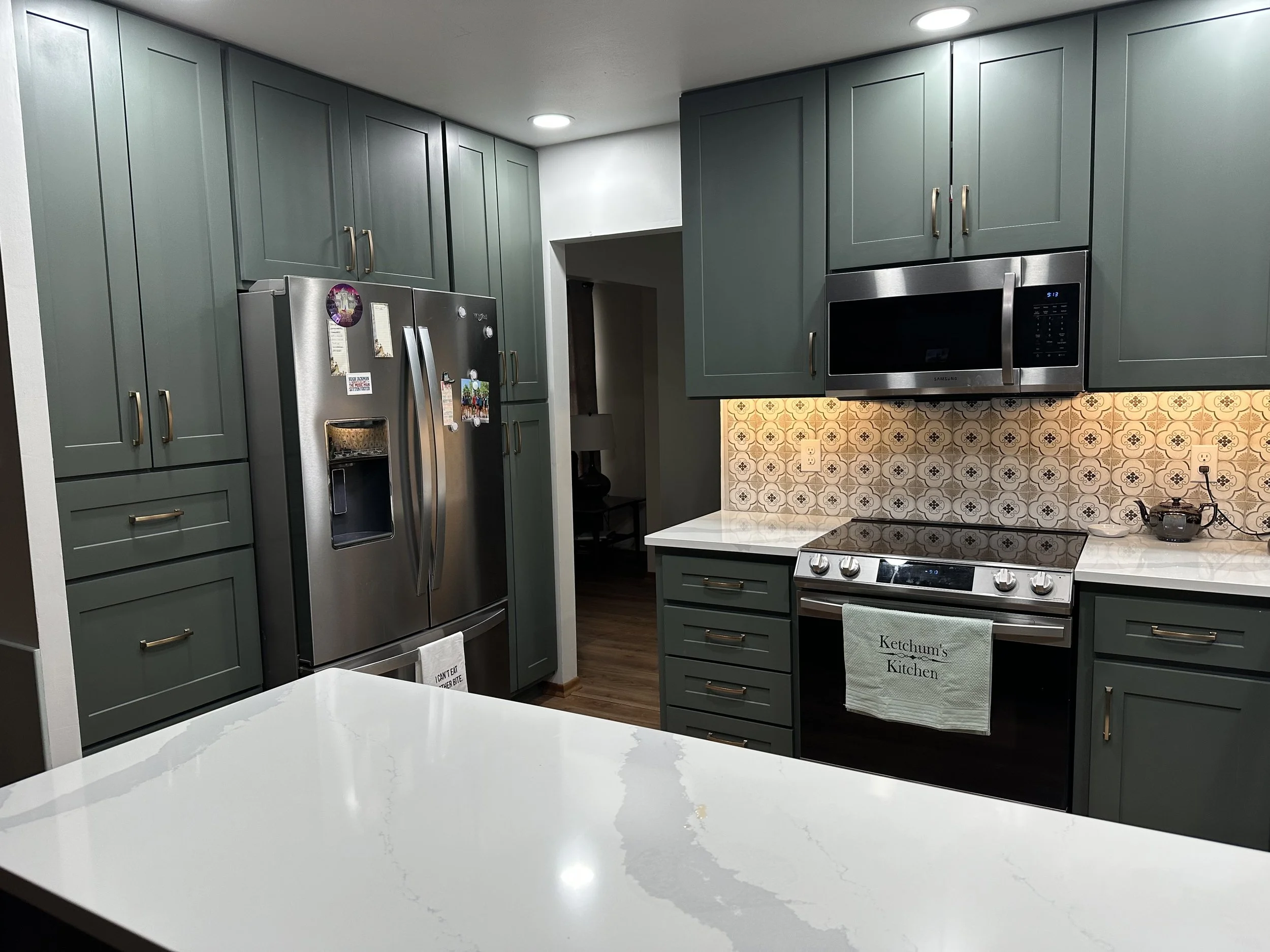 Modern kitchen with green cabinets, stainless steel appliances, patterned tile backsplash, white countertops, and a hanging towel labeled "Ketchum's Kitchen."