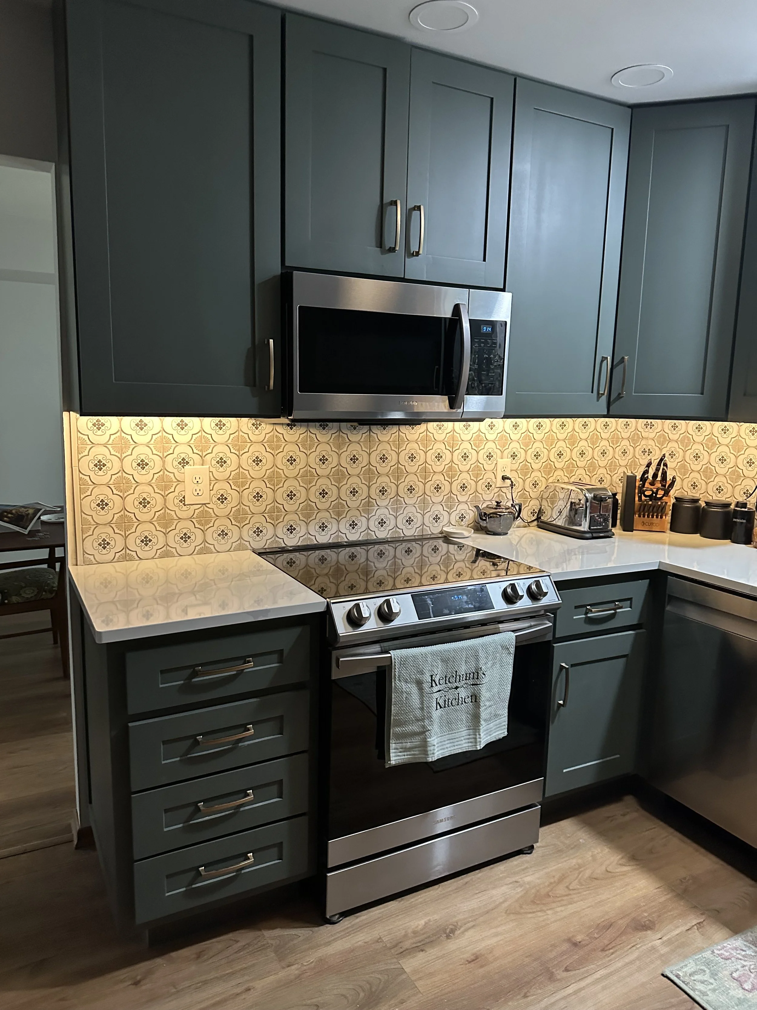 A kitchen with rich green cabinets, a tile backsplash with patterned design, and stainless steel appliances, including a microwave and oven. The countertop has small appliances and kitchen utensils.