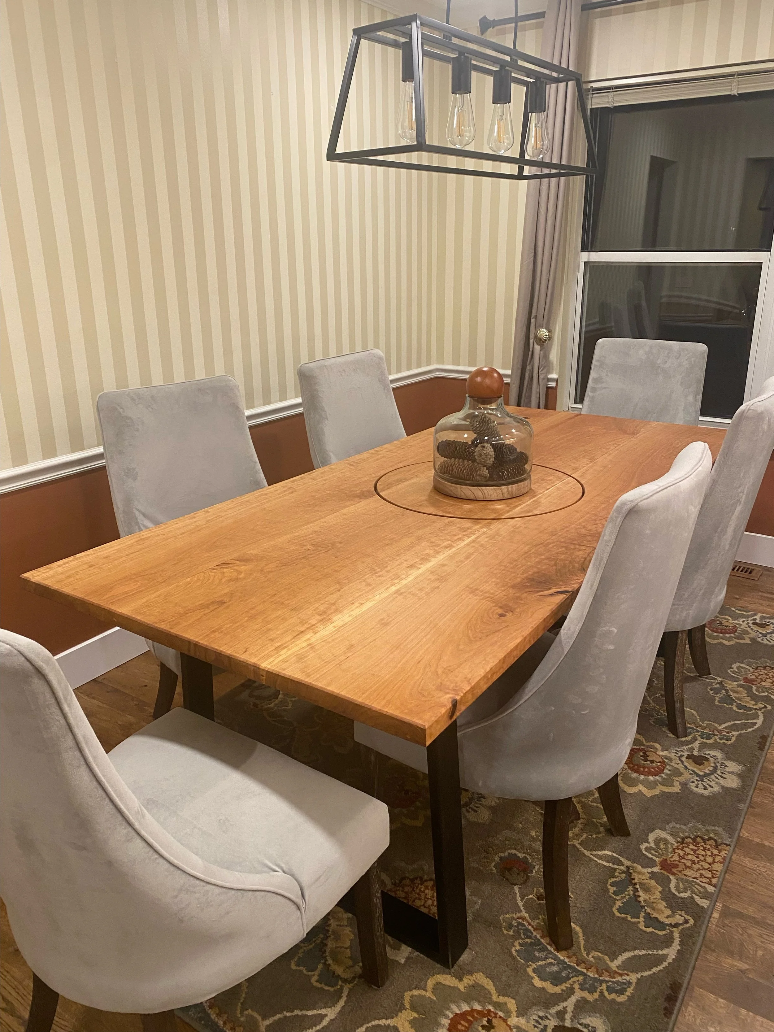 A wooden dining table with six upholstered chairs and a decorative glass jar filled with pinecones, a black metal chandelier with light bulbs hanging above, and a window with curtains in a dining room.