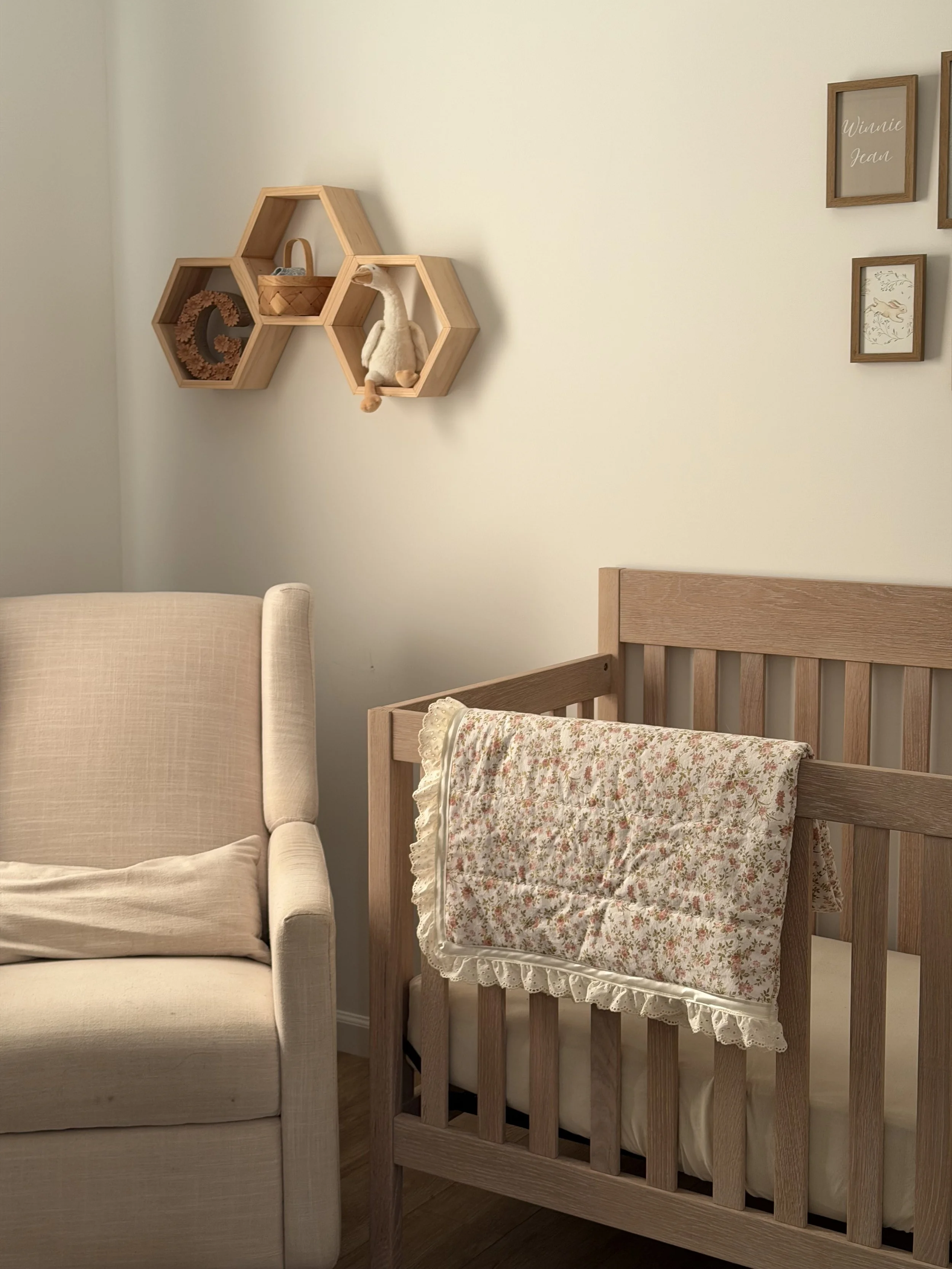 A room showing a beige armchair next to a wooden crib with a floral blanket hanging on it, and a set of wooden hexagonal wall shelves with decorative items.