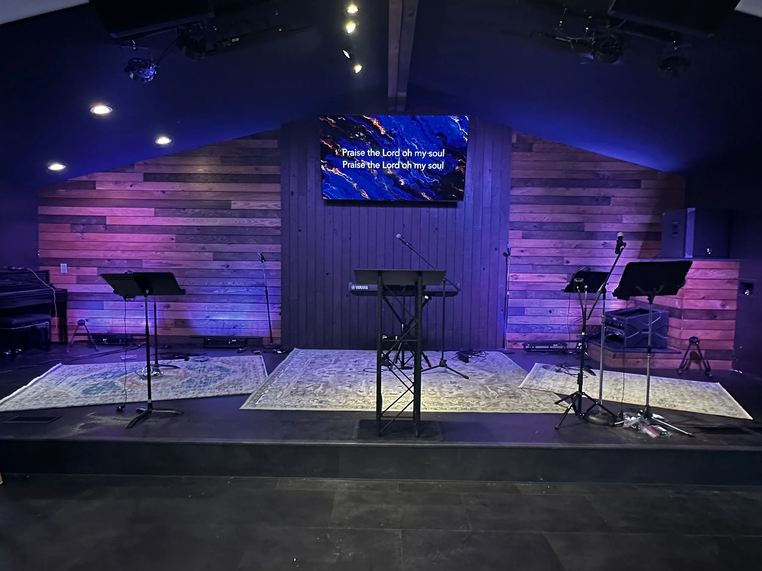 Empty stage in a church or worship center with musical instruments, microphones, and a large screen displaying song lyrics that read 'Praise the Lord oh my soul' against a blue, black, and orange abstract background.
