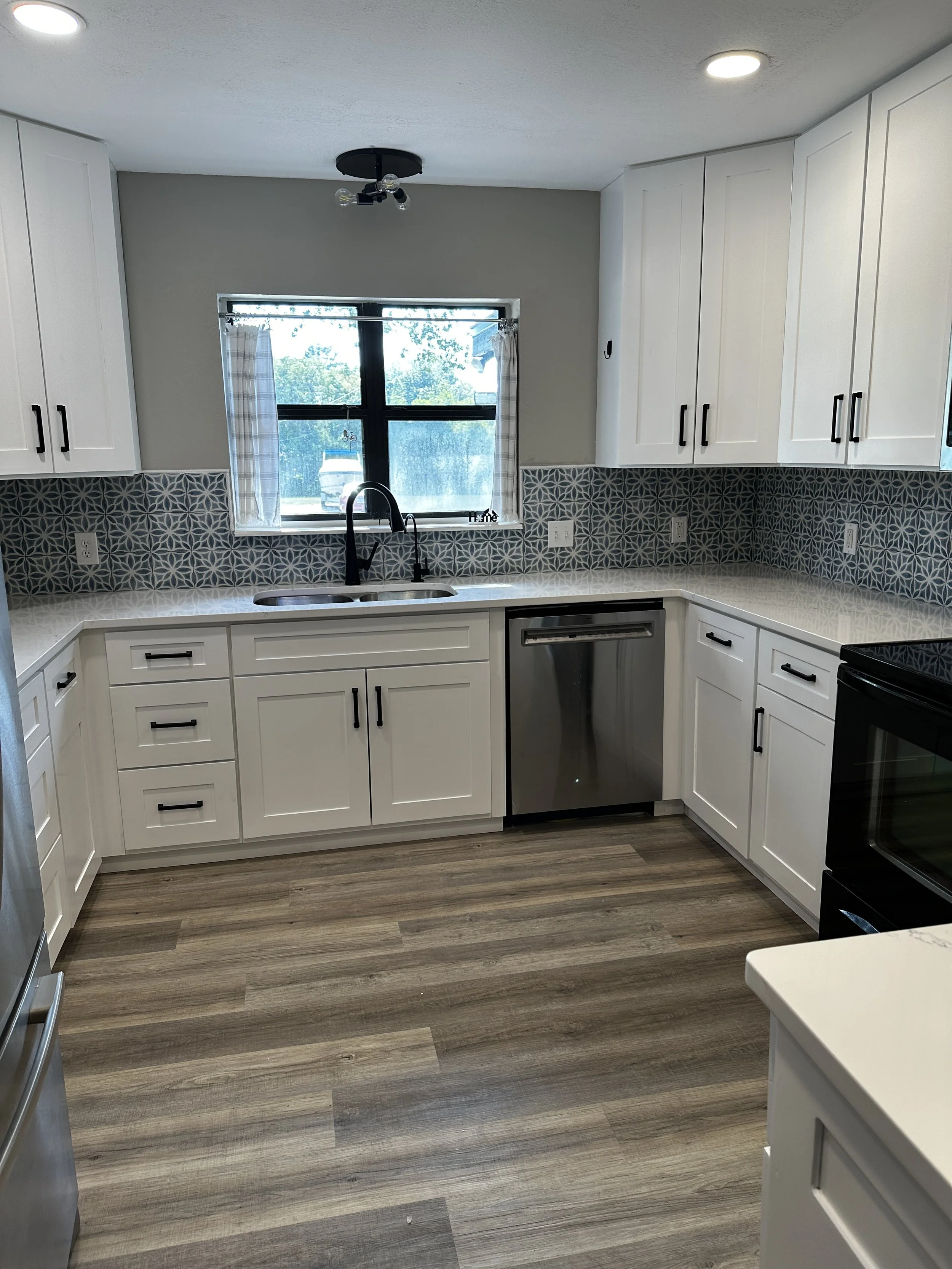 Modern kitchen with white cabinets, gray patterned backsplash, stainless steel dishwasher, black faucet, window with curtains, and wood-look flooring.