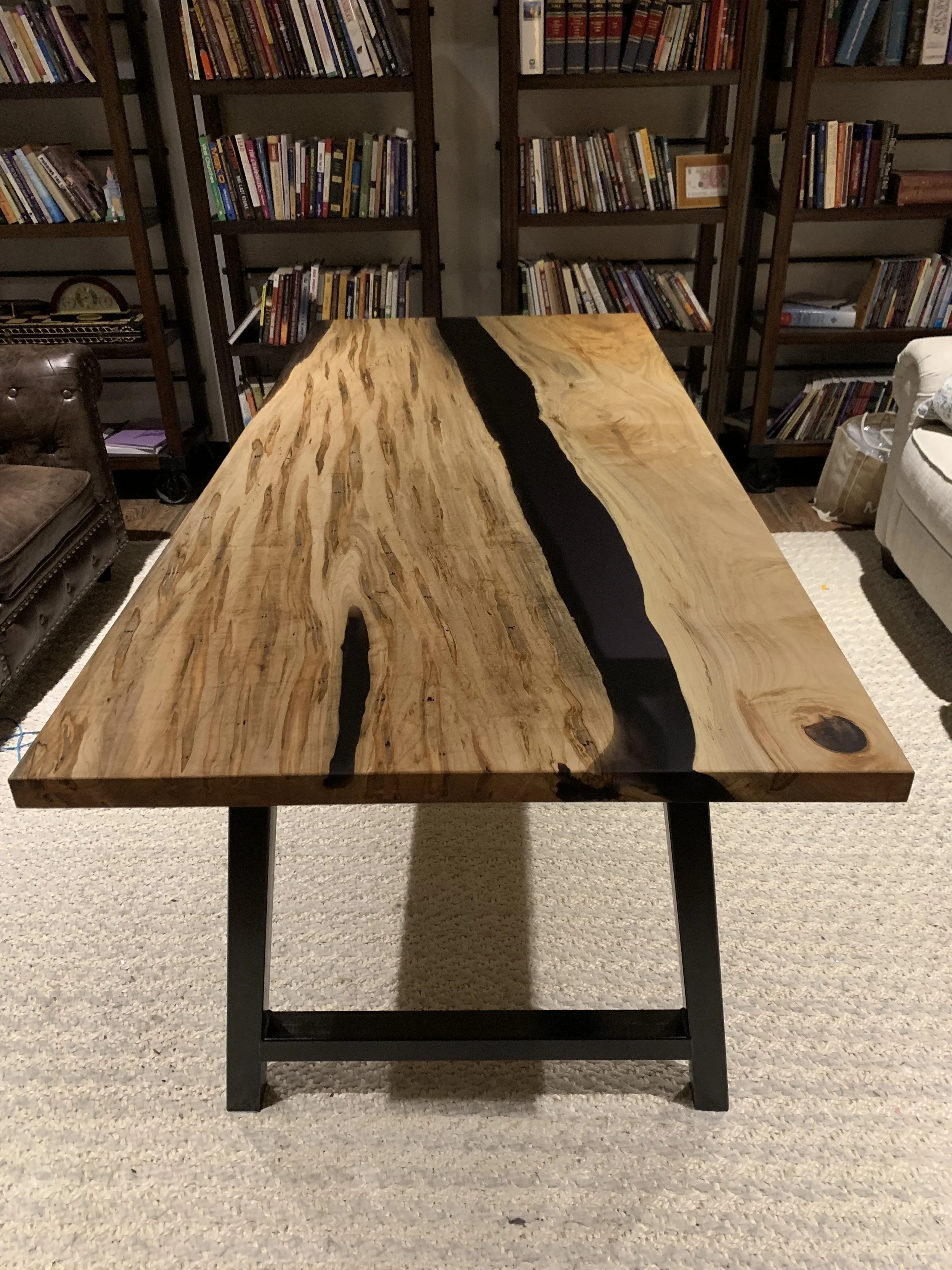 A wooden table with a natural, uneven edge and black resin inlay, in a room with bookshelves filled with books, a brown leather armchair, and a white upholstered chair.