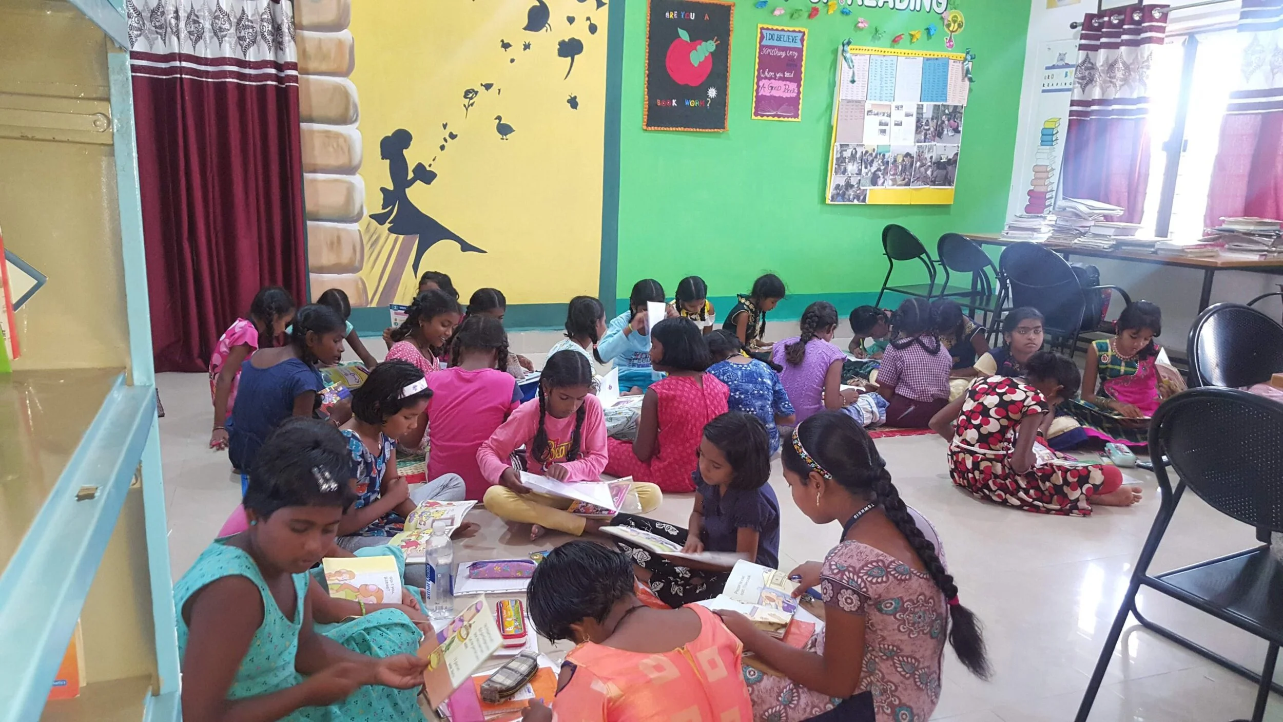A classroom filled with young girls sitting on the floor, reading and looking through books. The classroom has colorful walls with educational posters, student artwork, and stacks of papers on tables near the window.