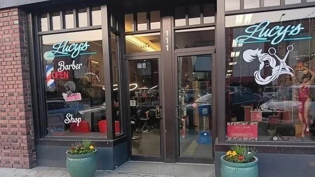 Fachada de un salón de belleza y barbería llamado Lucys, con ventanas grandes, unas plantas en macetas en la entrada, y un letrero que dice 'Open'.