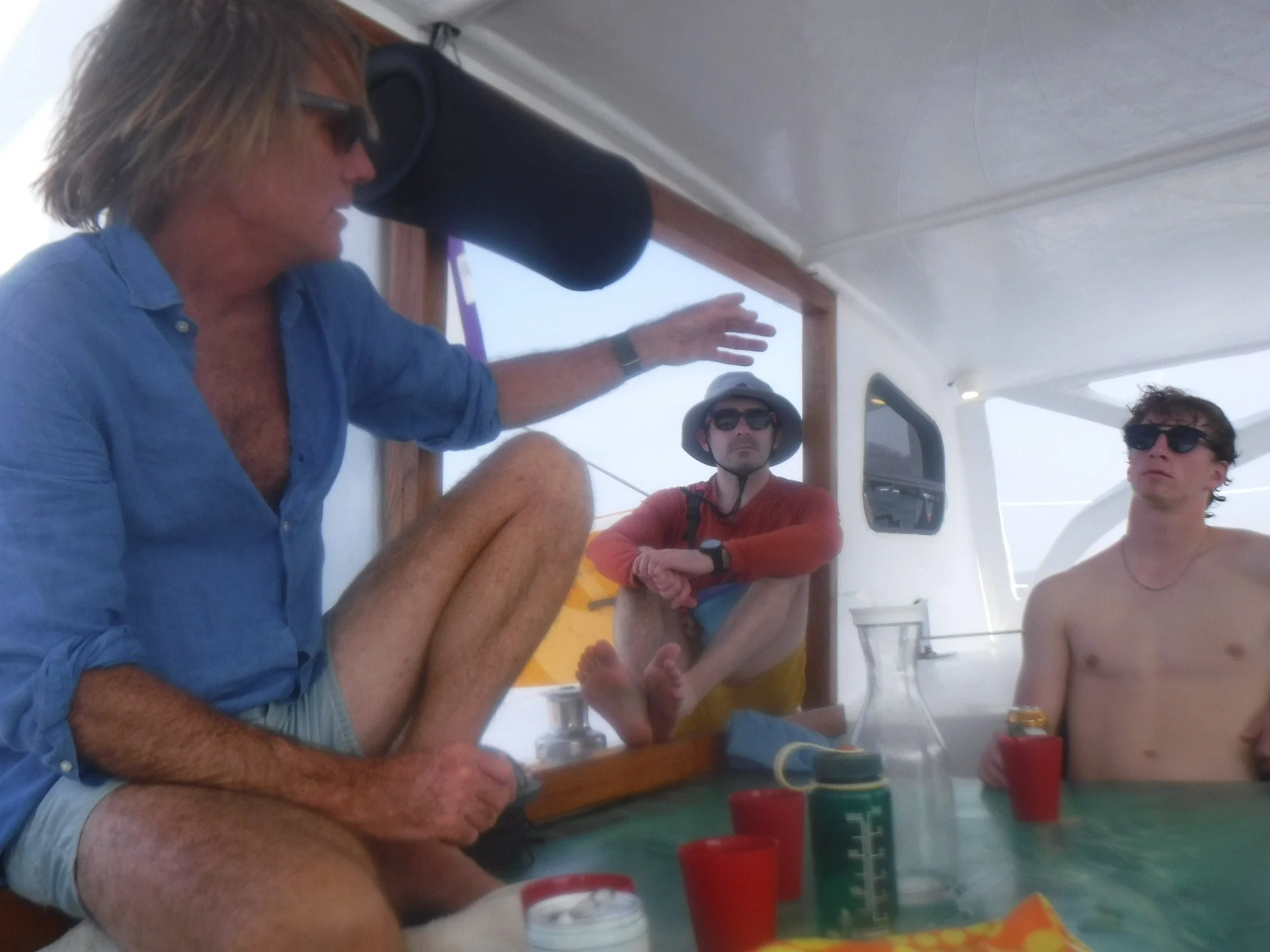 A group of people sitting at a table on a boat, engaged in conversation, with drinks and containers on the table.