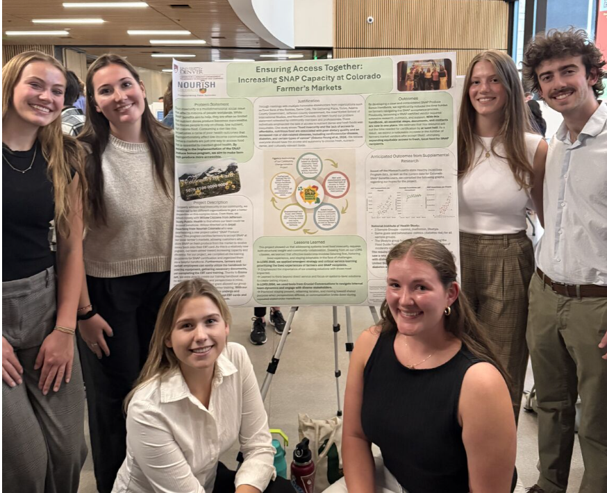 Group of six young people, three women and three men, presenting a scientific research poster at an academic event. Four of them are standing, two are kneeling in front, all smiling and looking at the camera. The poster is titled 'Ensuring Access Together: Increasing SNAP Capacity at Colorado Farmer's Markets' and includes various sections, charts, and images.
