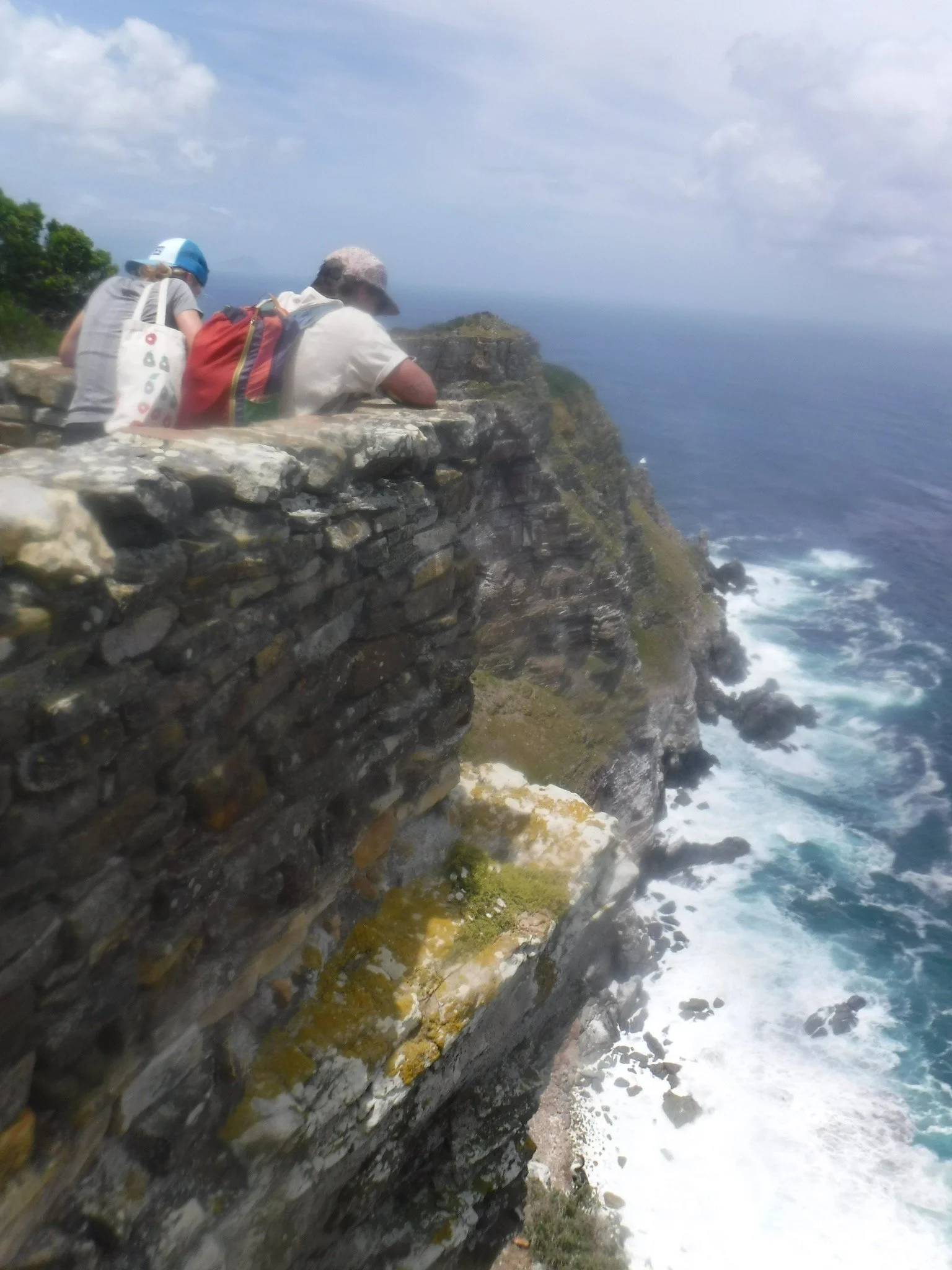 People sitting on a stone wall overlooking a rugged coastline with cliffs and the ocean.